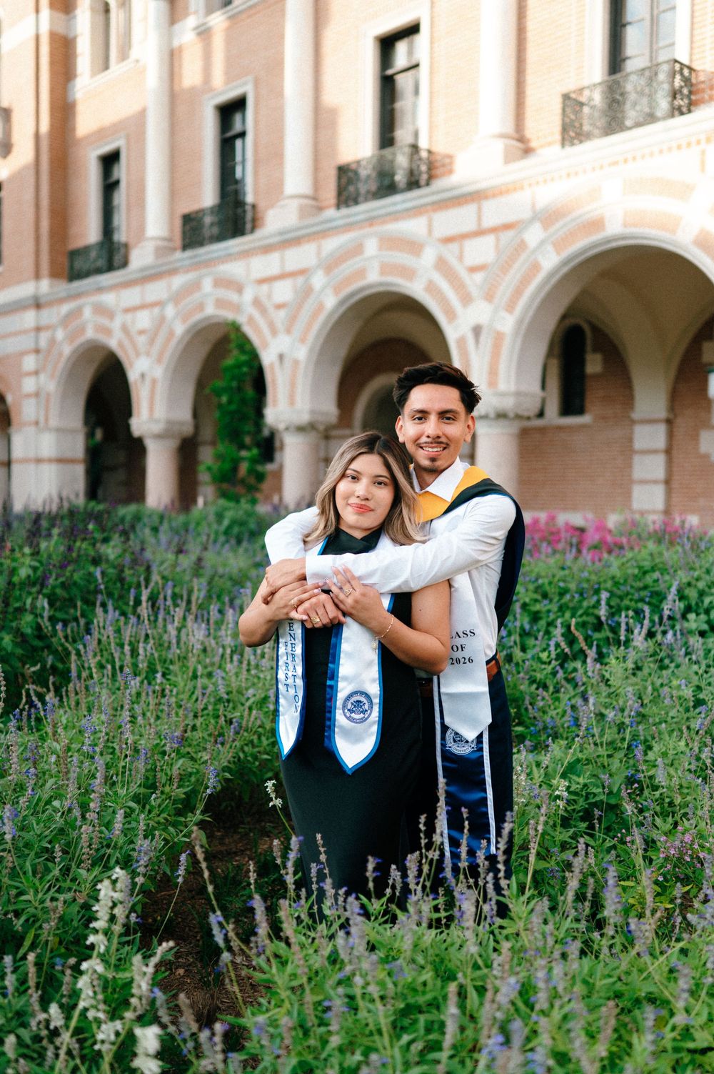 Two graduates in black gowns and stoles embrace in a garden courtyard of a university building.