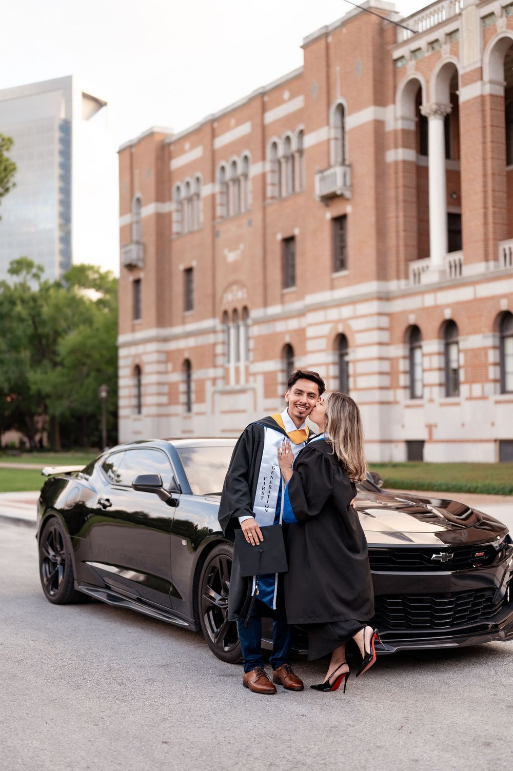Two graduates in black robes pose in front of a black Chevrolet Camaro at a brick university building.