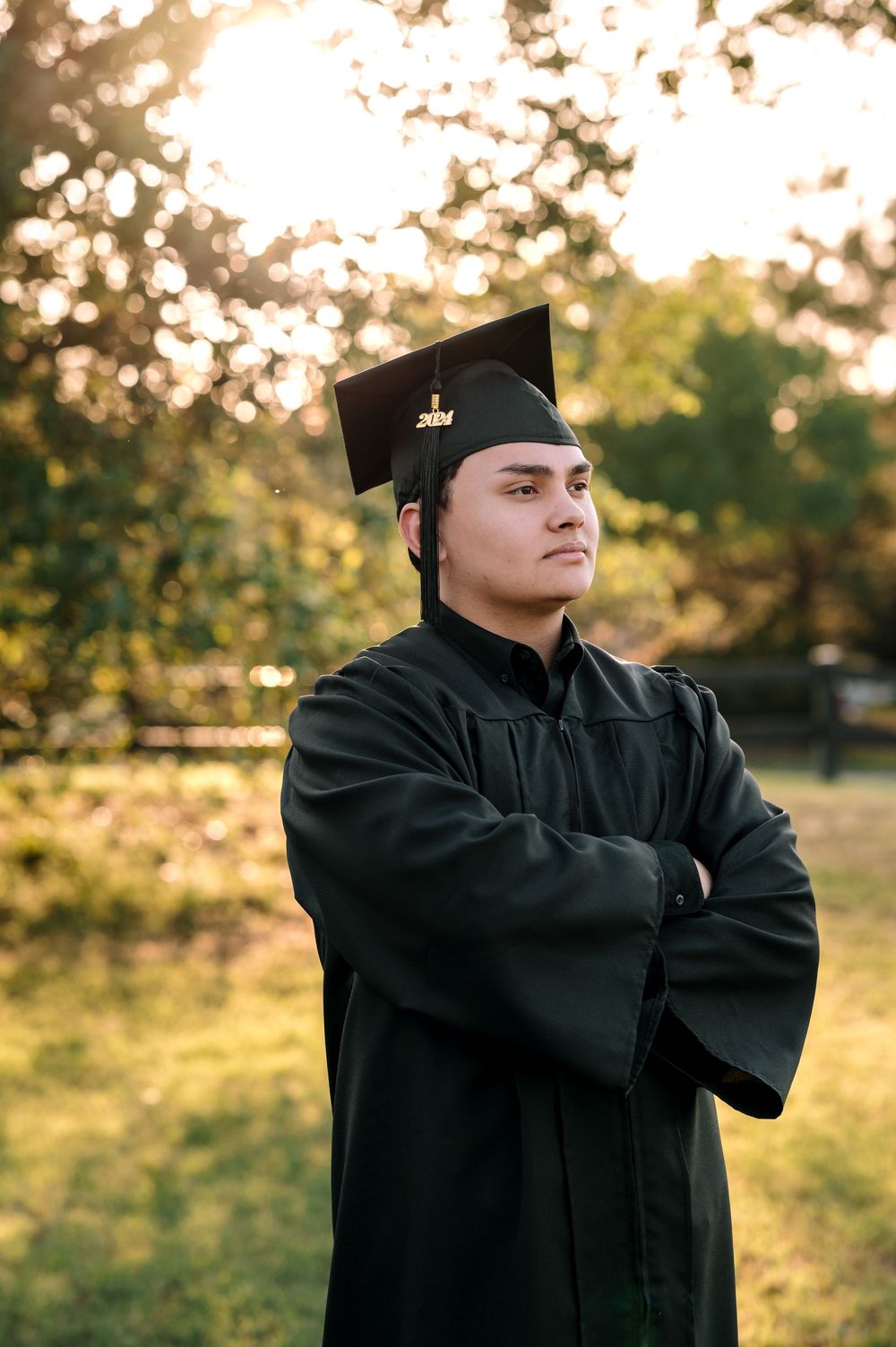 Graduate in traditional black academic regalia poses outdoors at sunset with arms crossed.