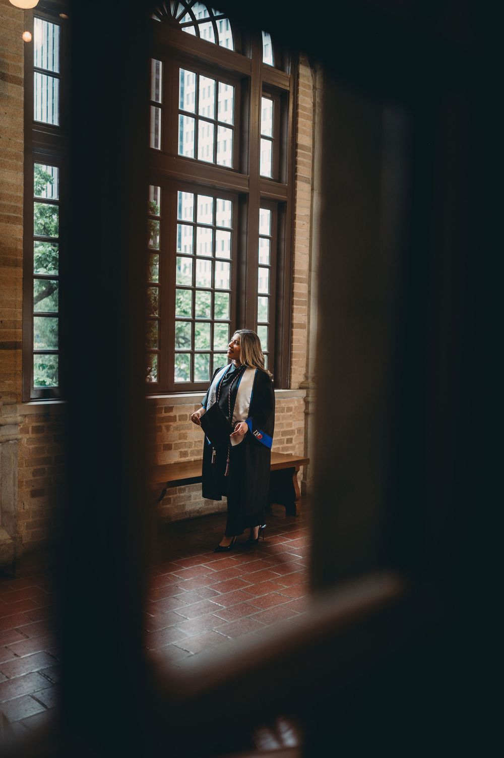 Graduate in academic regalia stands by tall windows in a historic brick building, holding diploma during graduation.