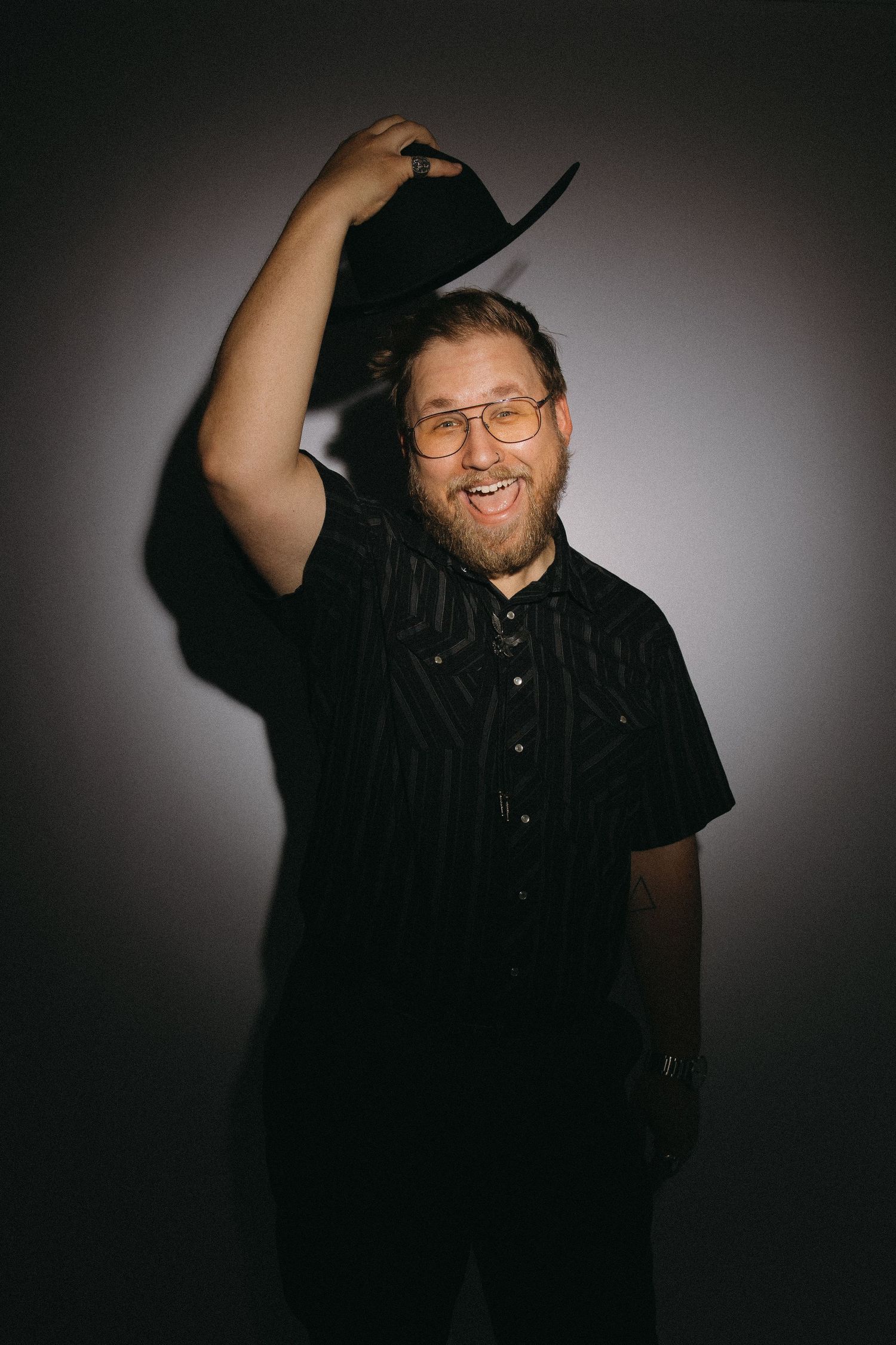 Portrait of a person in black shirt raising their hat while smiling against dark background.