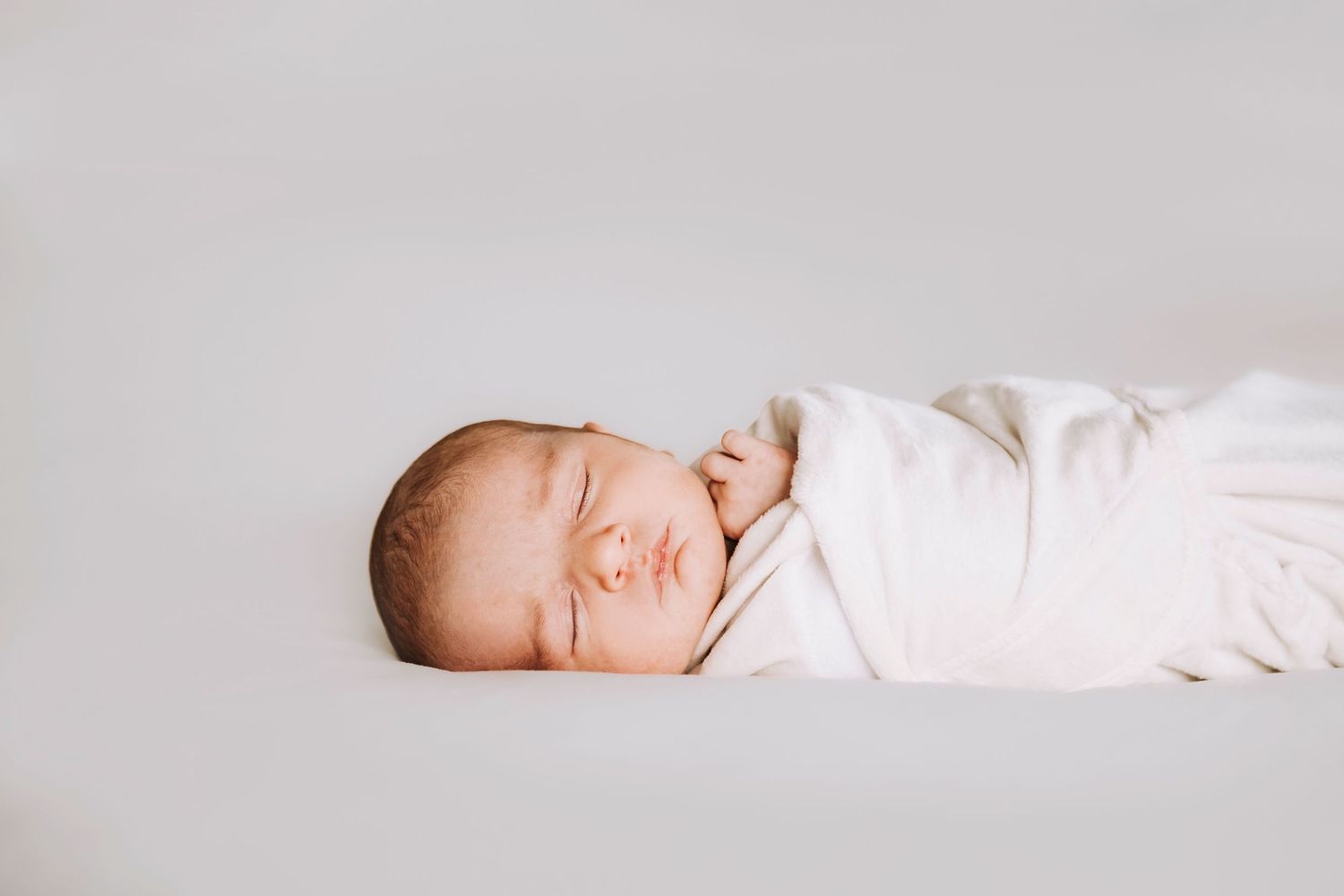 Serene newborn baby sleeping peacefully wrapped in white against light gray backdrop.
