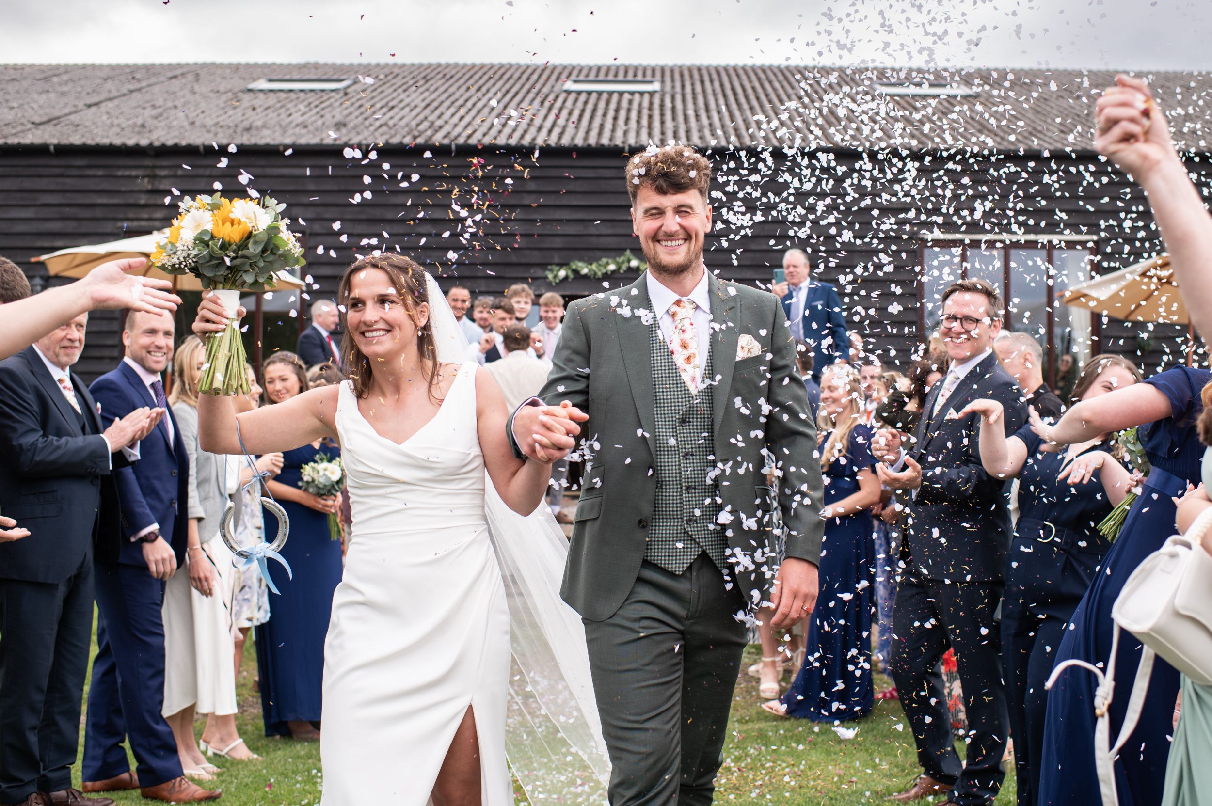 Bride and groom walk towards the camera - they are smiling, the groom has closed his eyes as white paper heart confetti is thrown by guests