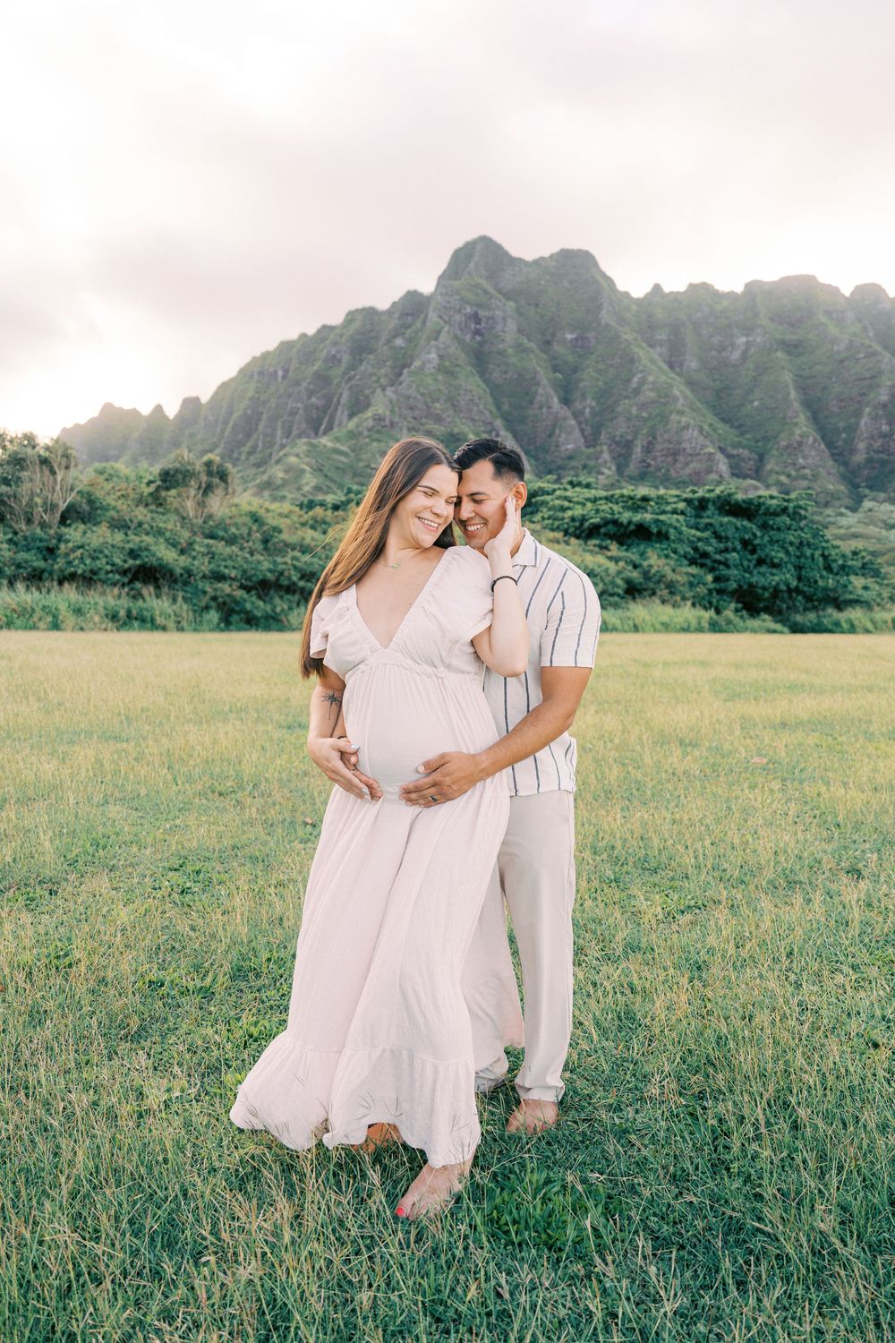 Couple embracing in green meadow with dramatic Ko'olau Mountains backdrop during maternity photo session in Hawaii.