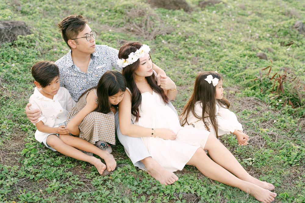 A family of five sits together on green grass, with a pregnant woman wearing a floral crown in a white dress.