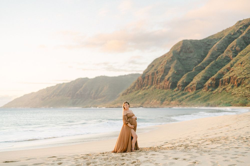 Pregnant woman in flowing golden gown poses on sandy Hawaiian beach with dramatic green mountains and ocean at sunset.