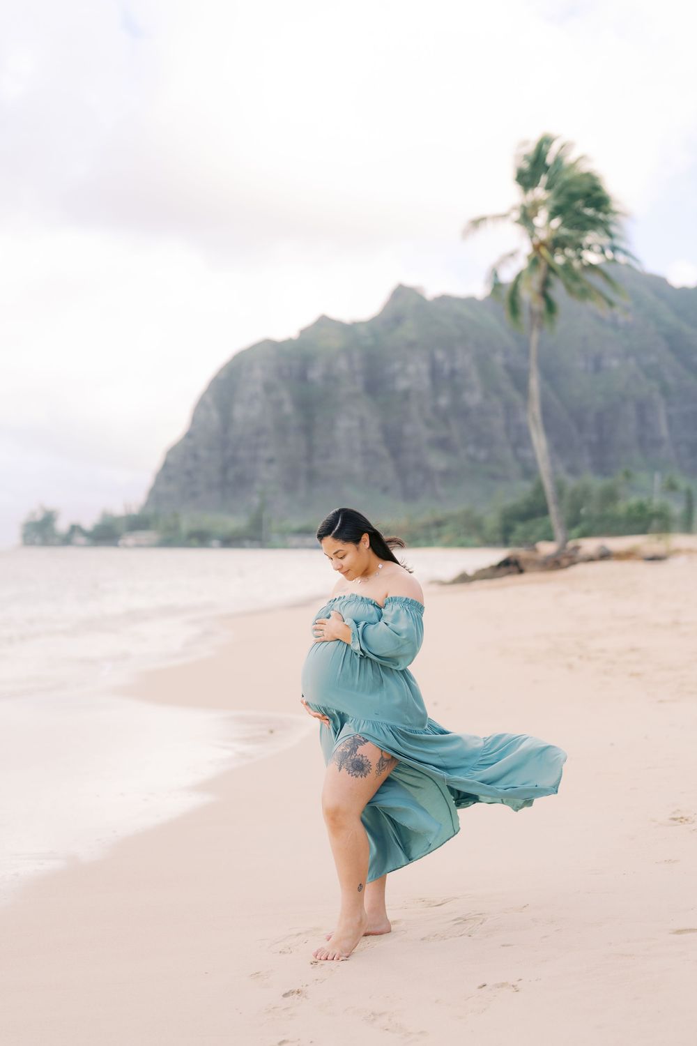 Pregnant woman in teal dress on Hawaiian beach with mountain and palm tree backdrop.