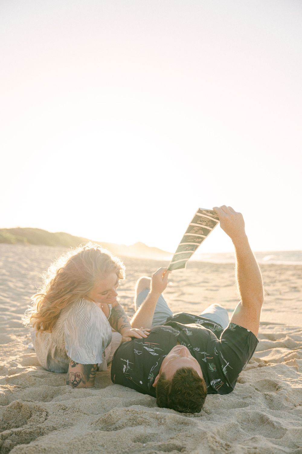 Couple relaxing on sandy beach at sunset, woman leaning over man lying down reading a book together.