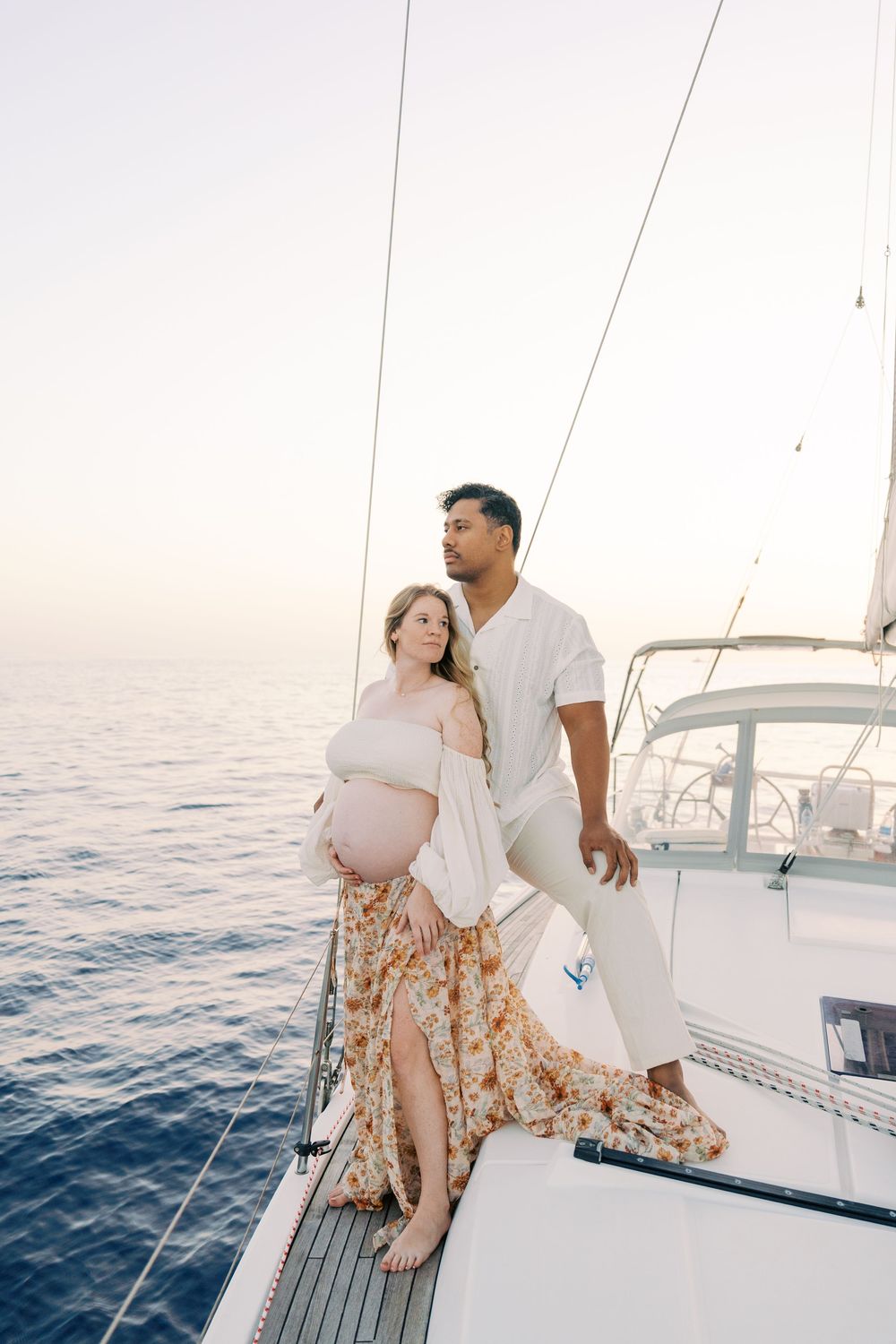 Pregnant couple embracing on a sailboat deck at sunset, with calm blue ocean waters in the background.