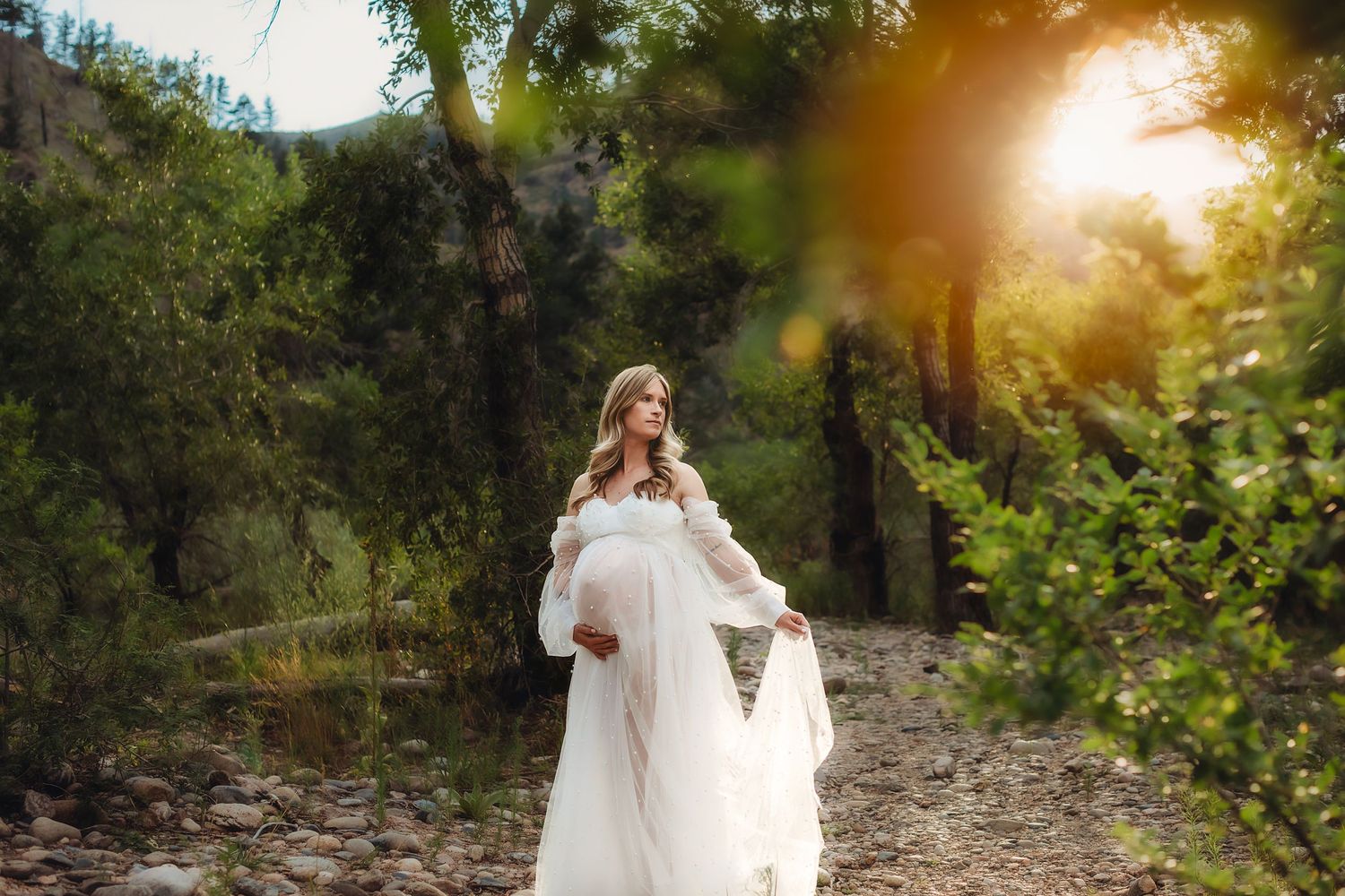 Woman in flowing white dress stands in sunlit forest setting during maternity photoshoot.