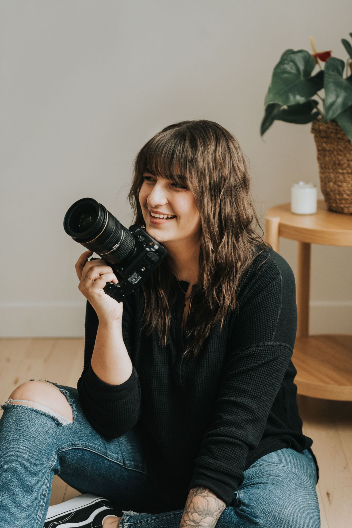 A photographer in a black sweater and ripped jeans sits on the floor holding a professional camera while smiling.