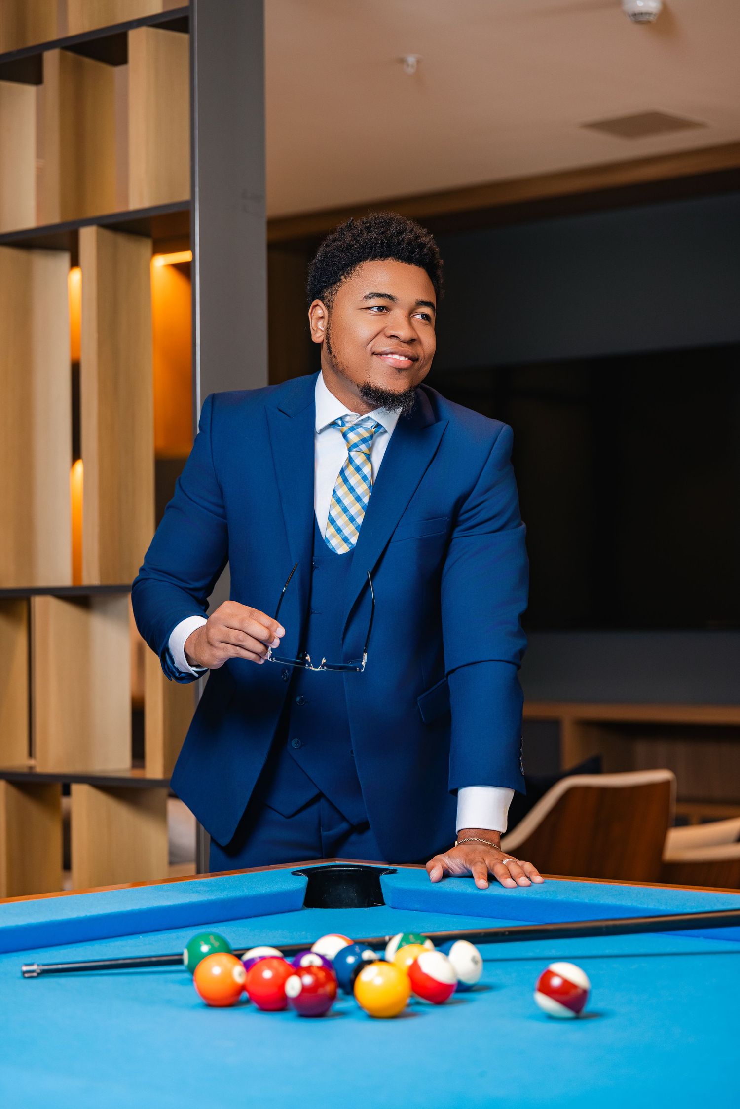 Confident man in blue suit stands beside a pool table holding glasses in a modern lounge setting.
