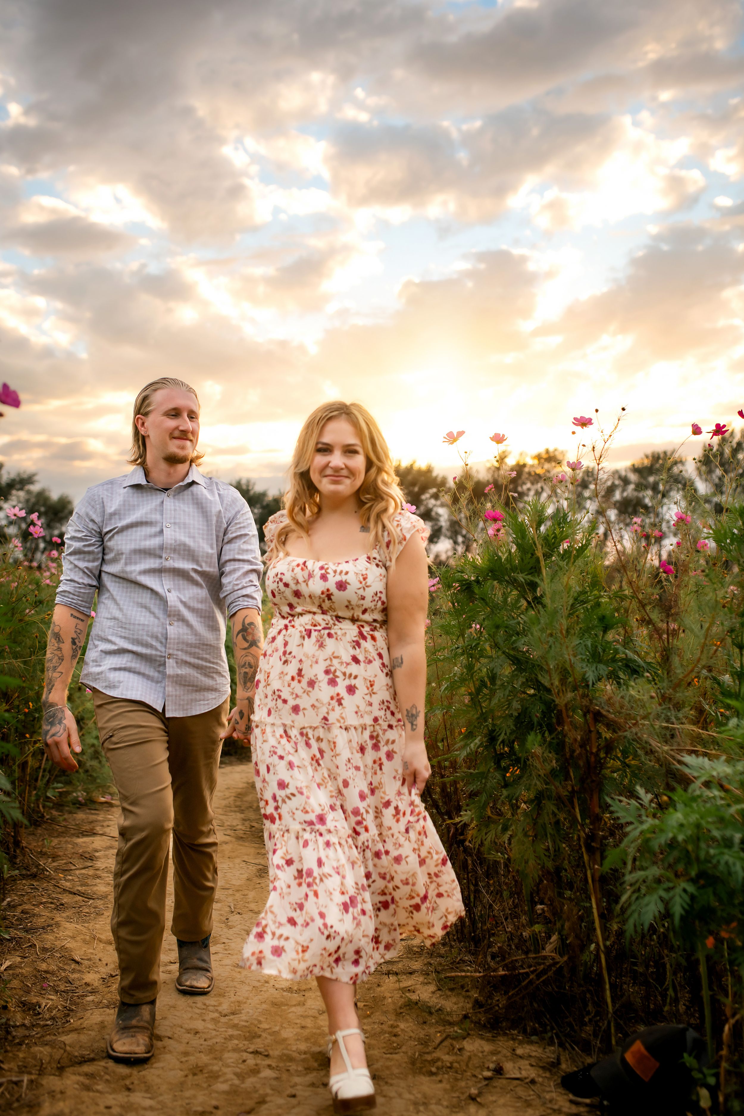 A young couple walks together through a field of flowers at sunset, with a cloudy sky in the background.