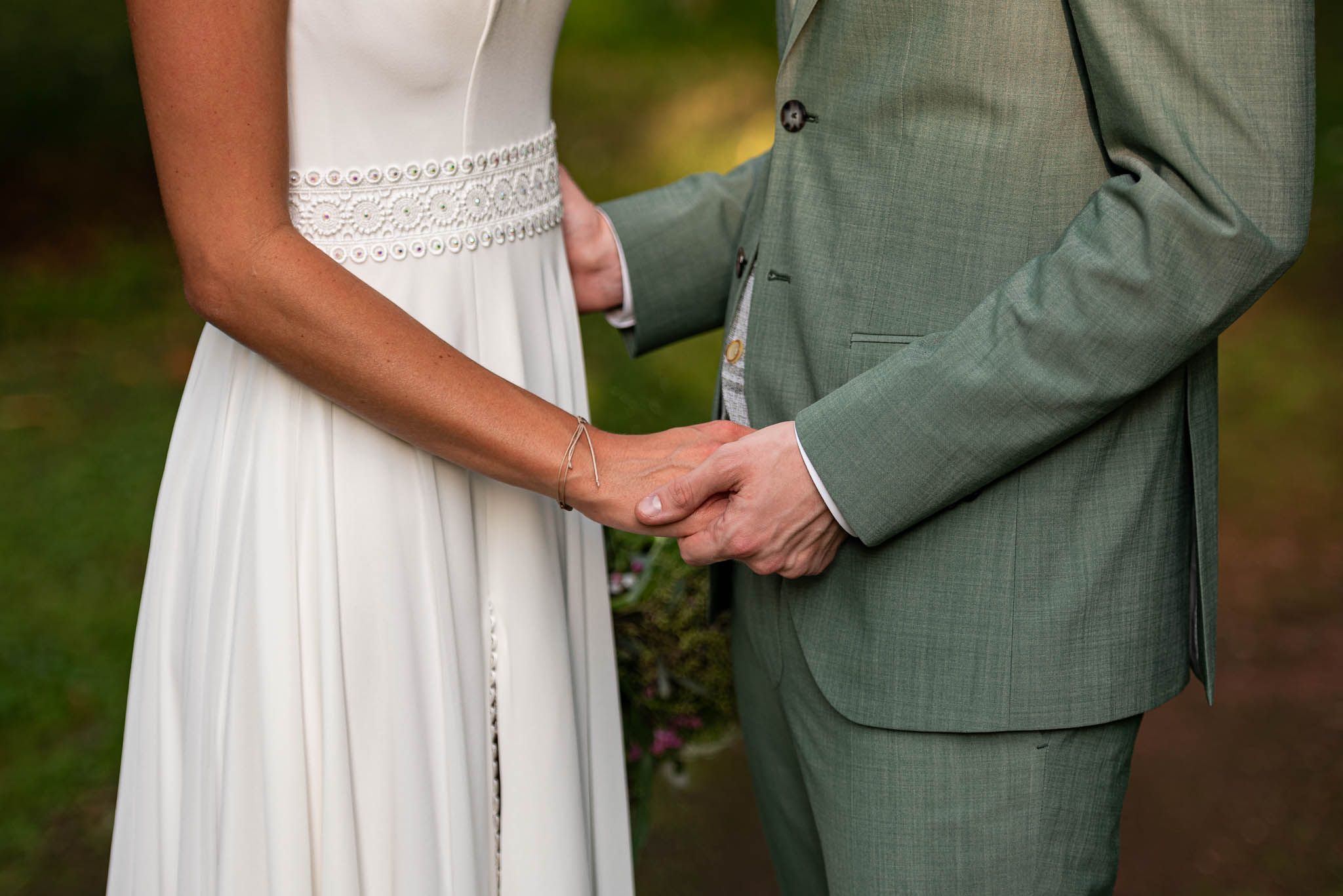 Nahaufnahme von Händen, die sich zwischen einem weißen Hochzeitskleid und einem salbeigrünen Anzug während der Freiluftzeremonie halten.