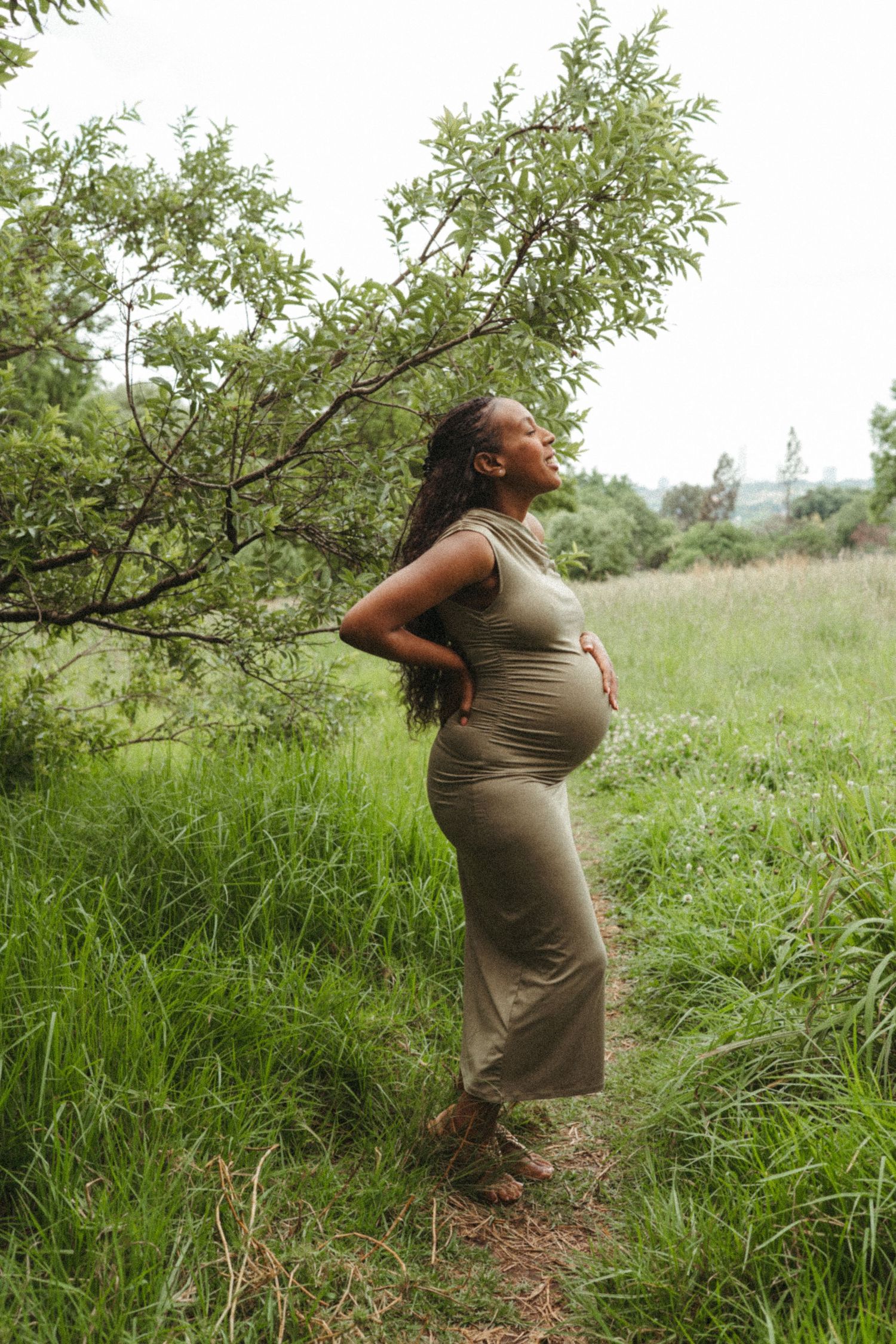 A pregnant woman in a beige dress stands in a lush green field with trees in the background.
