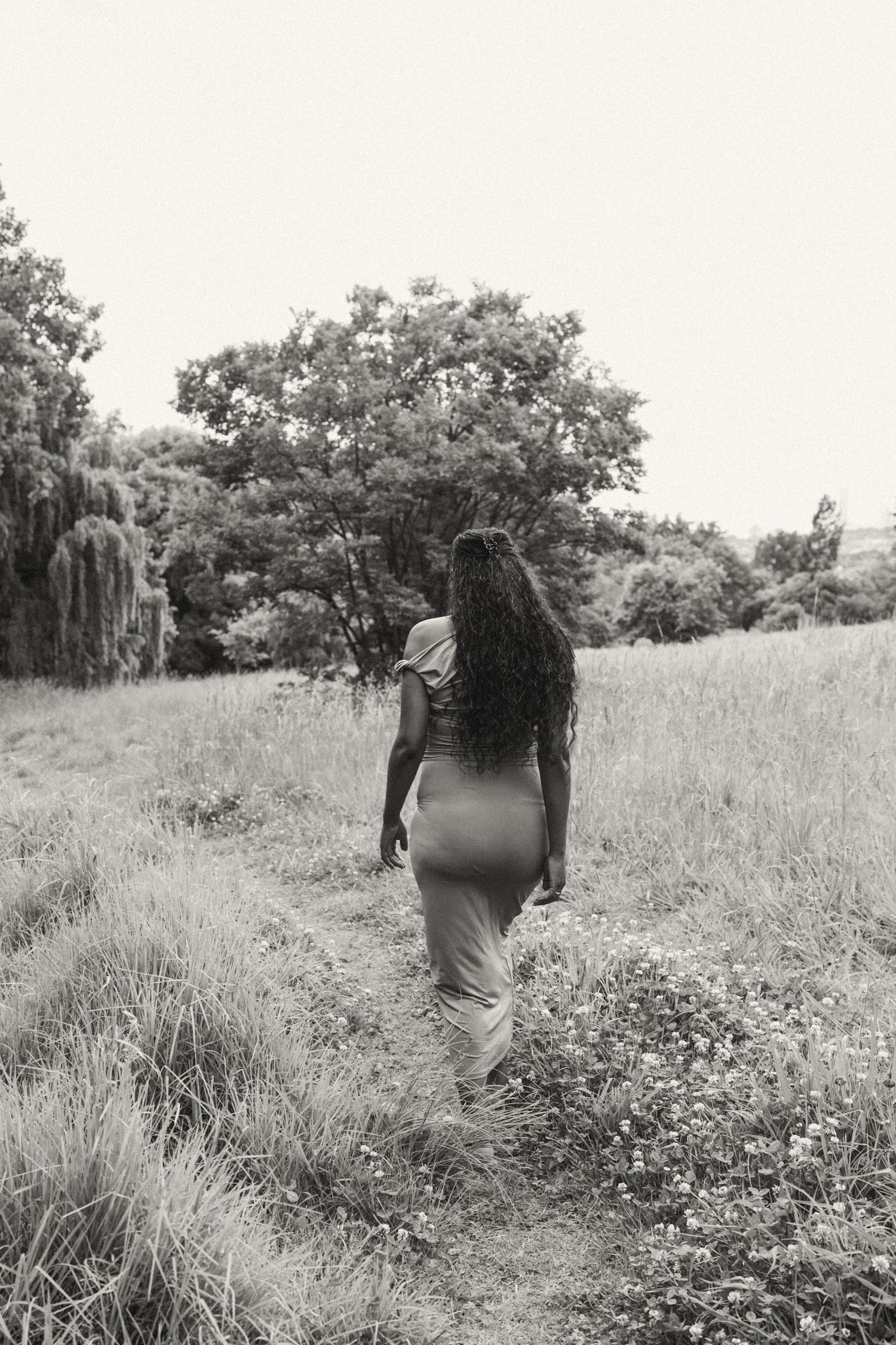 Black and white photo of a person walking through a flowering meadow with trees in the distance.