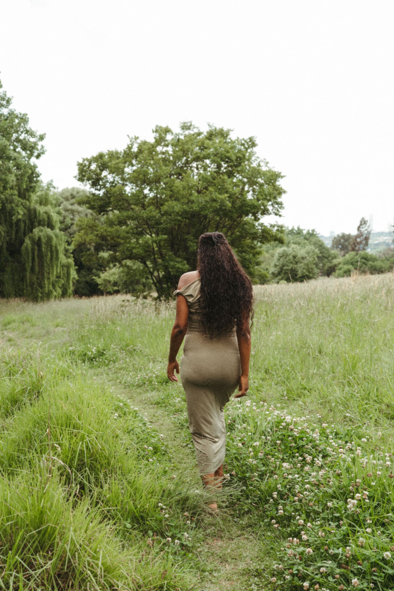 Woman in a tan dress walks through a wildflower meadow surrounded by green trees.