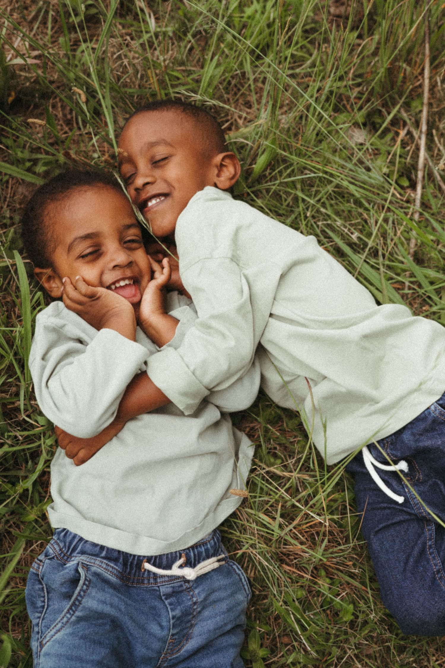 Two young children in matching sweaters lying in grass and laughing together.