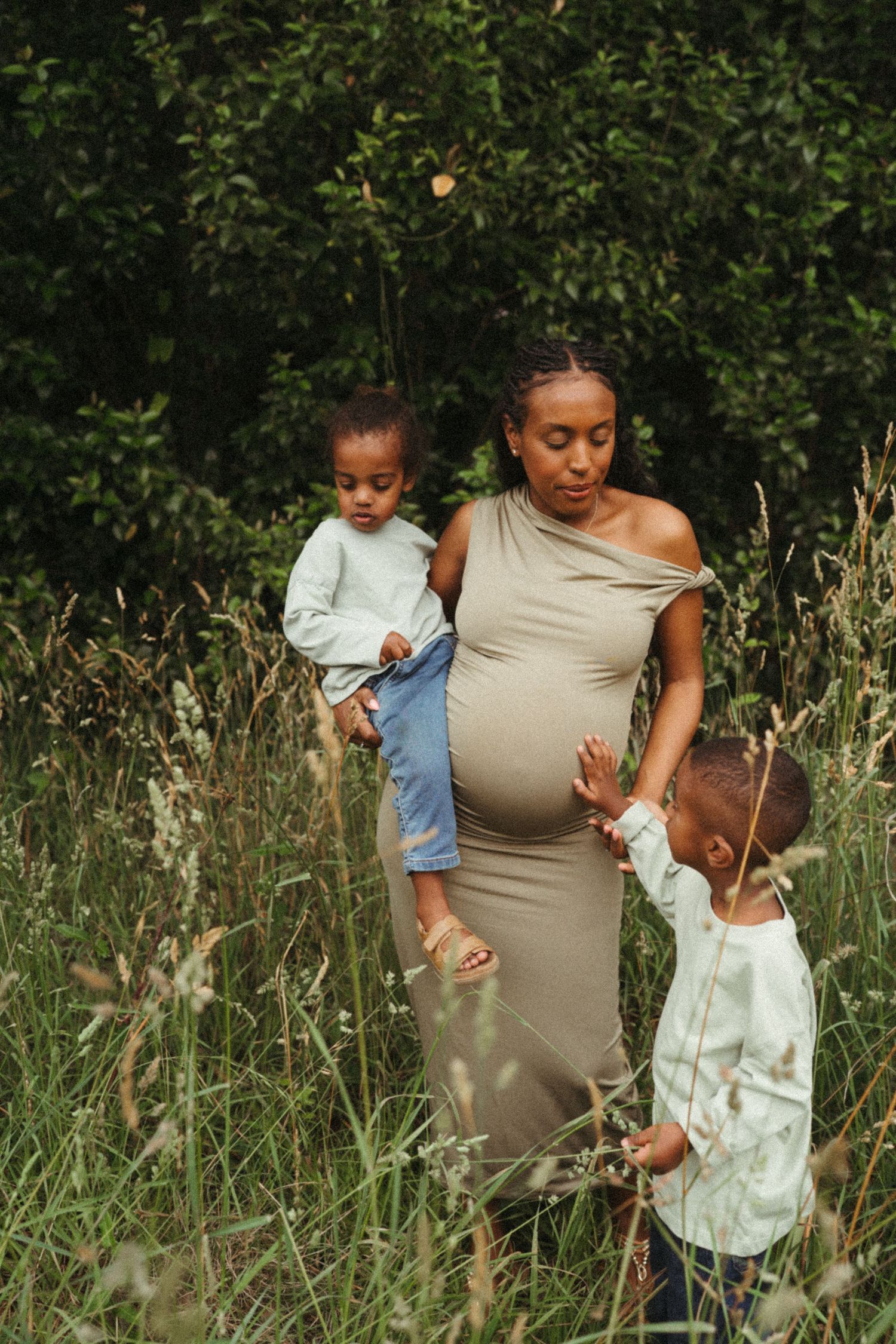 Expectant mother in beige dress standing with children in a lush garden setting.