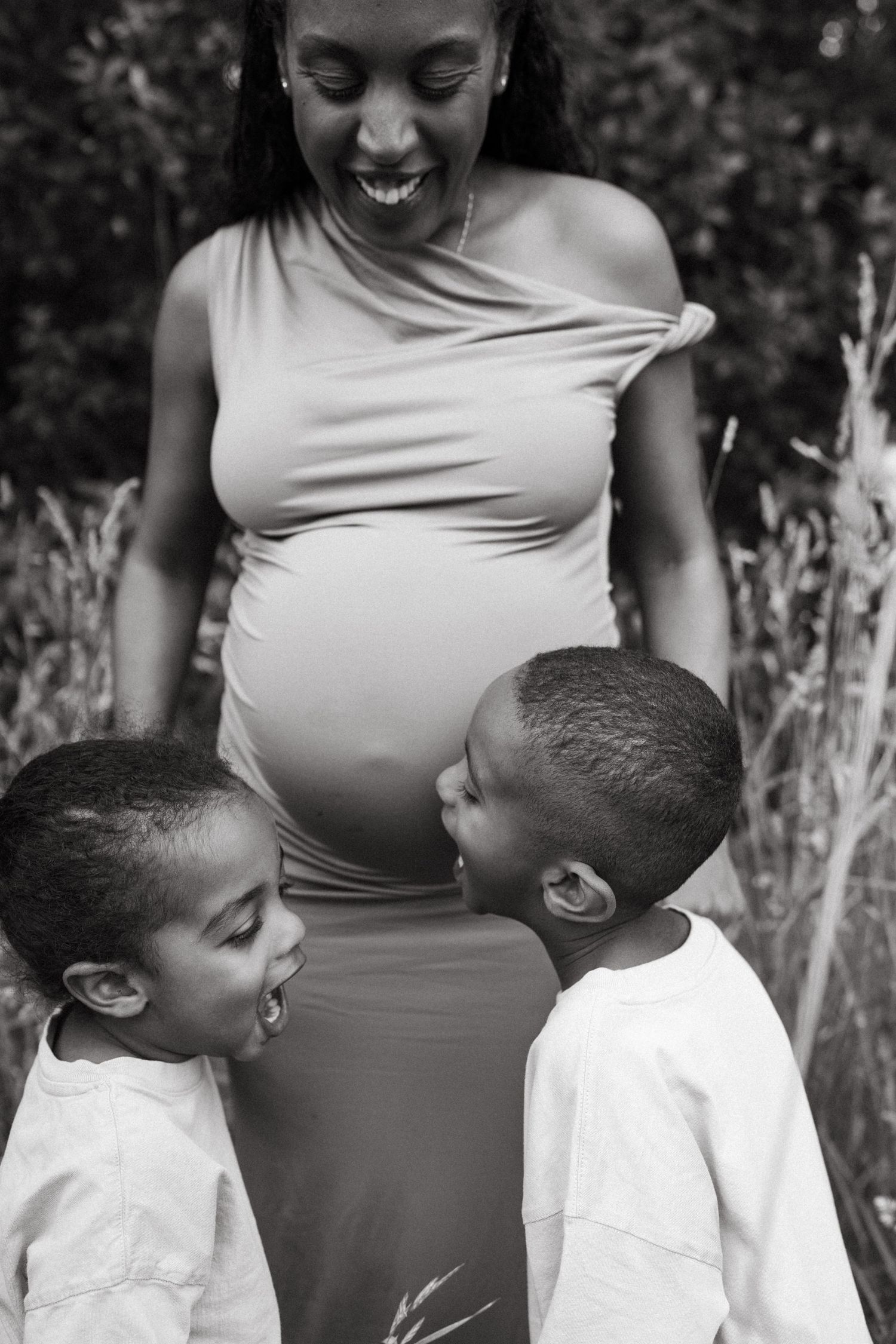 Black and white portrait of an expectant mother sharing a moment with children in tall grass.