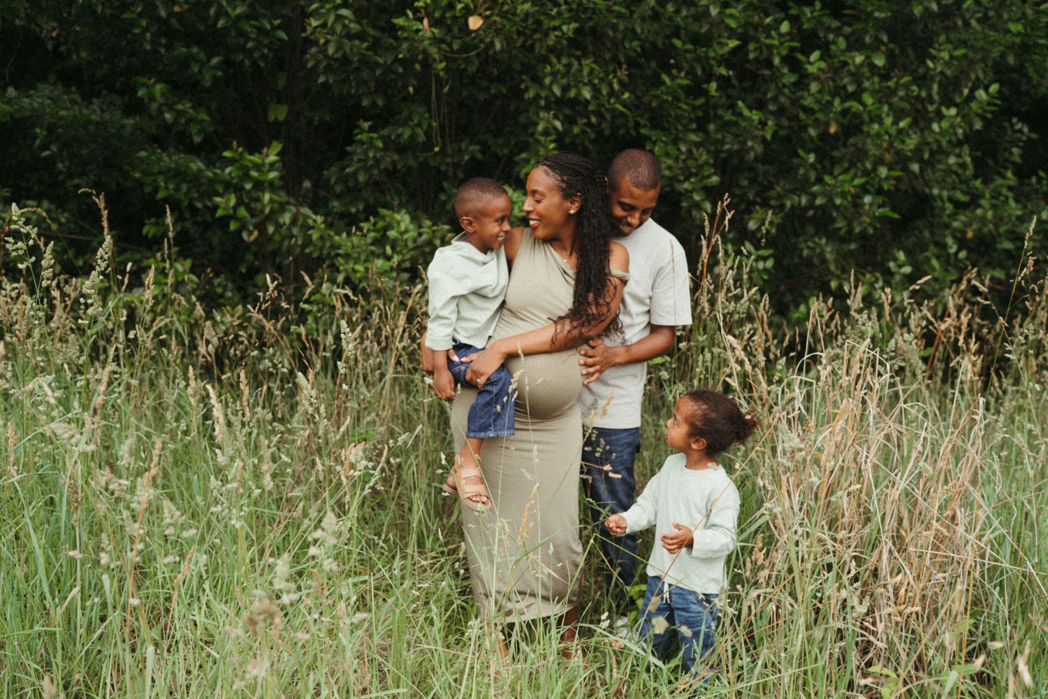 Family enjoying a tender moment together in a sunlit meadow surrounded by greenery.