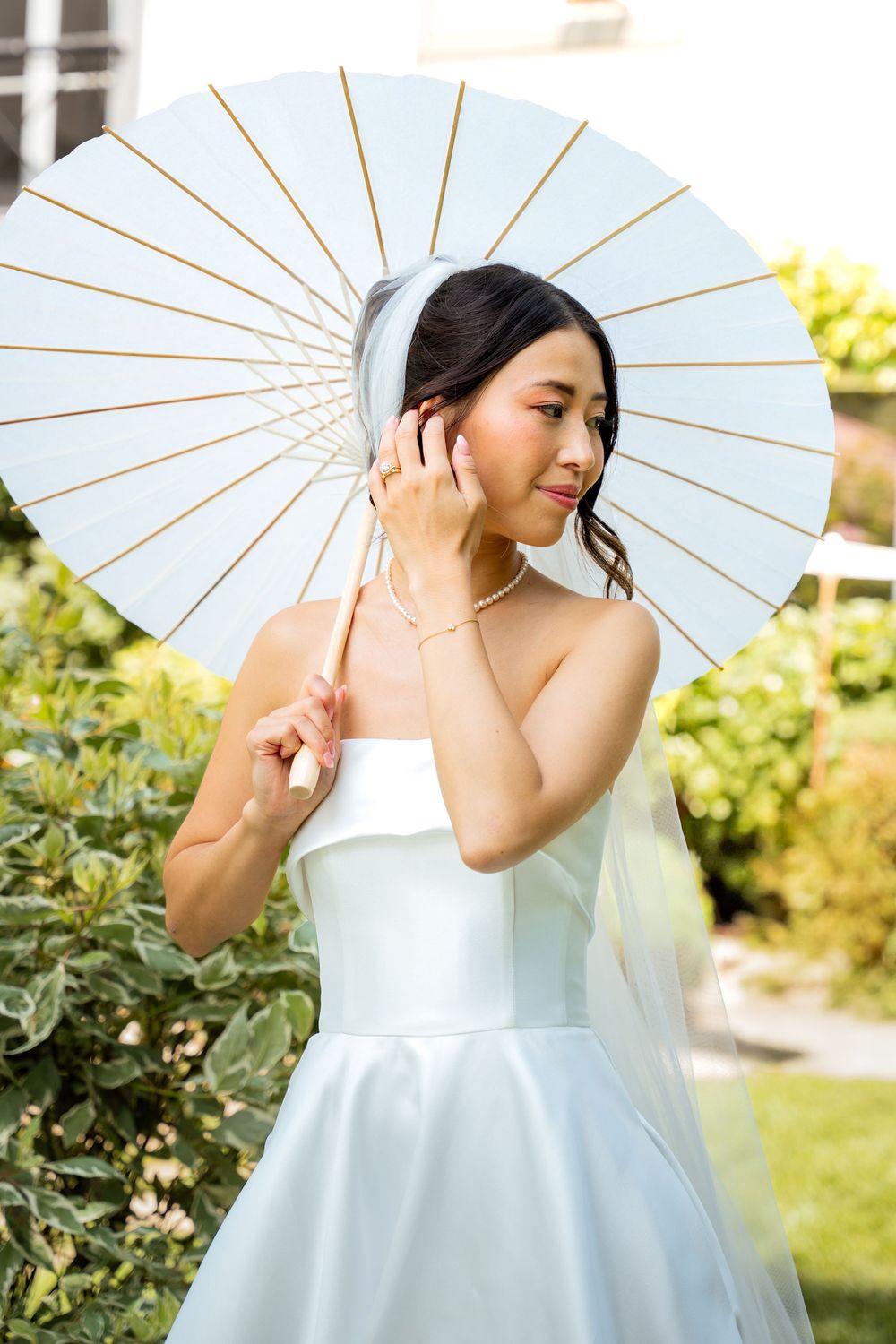 Bride in elegant white dress poses with traditional paper parasol in garden setting.
