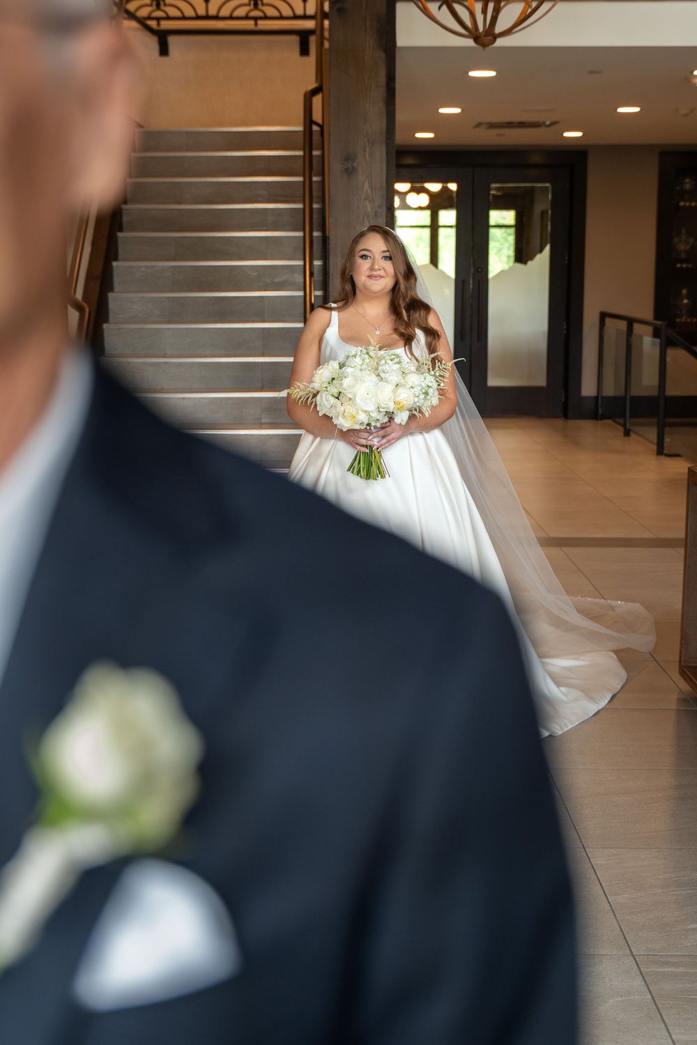 A bride in a white wedding gown holds a bouquet while walking through an elegant lobby with dark wood accents.