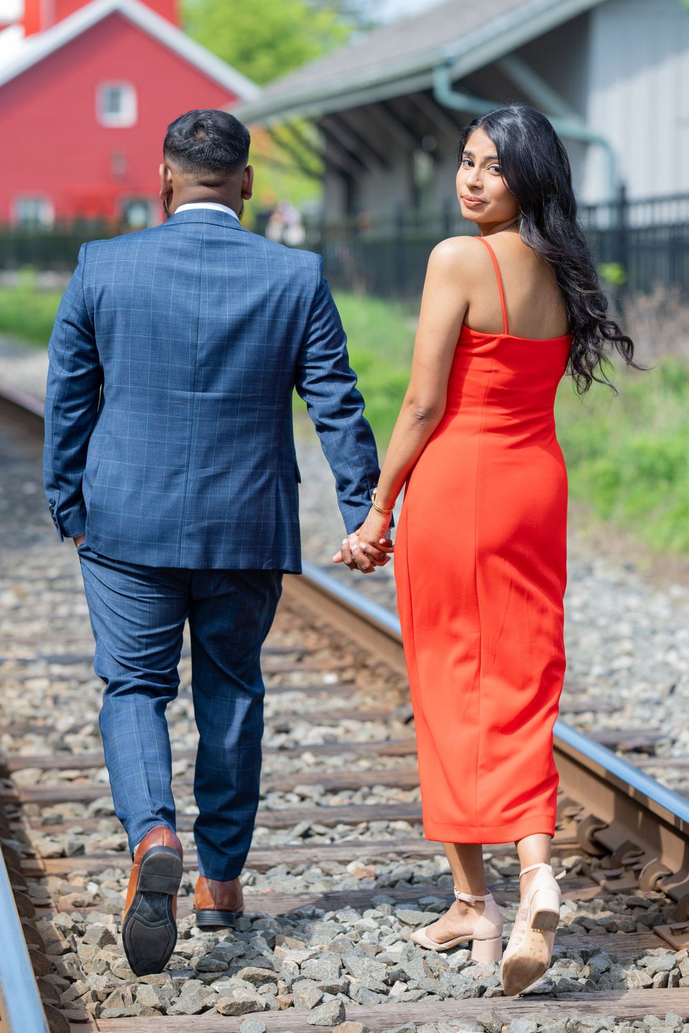 Couple walking hand in hand along railroad tracks with red building in background.