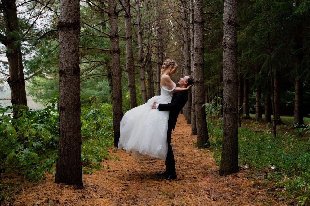 Romantic woodland photo of groom lifting bride in white dress among pine trees.