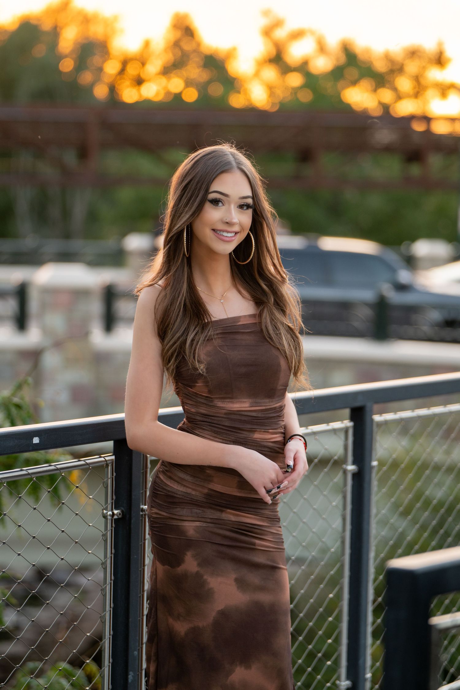 High school senior in brown satin dress poses on outdoor deck during golden hour.