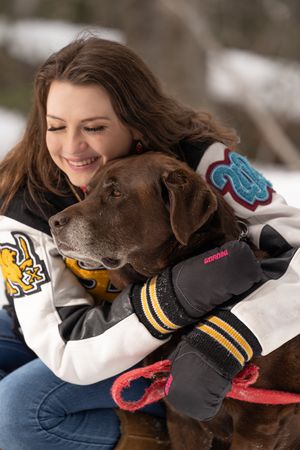 A chocolate labrador wearing a sports jersey playfully interacts with someone in a winter setting.