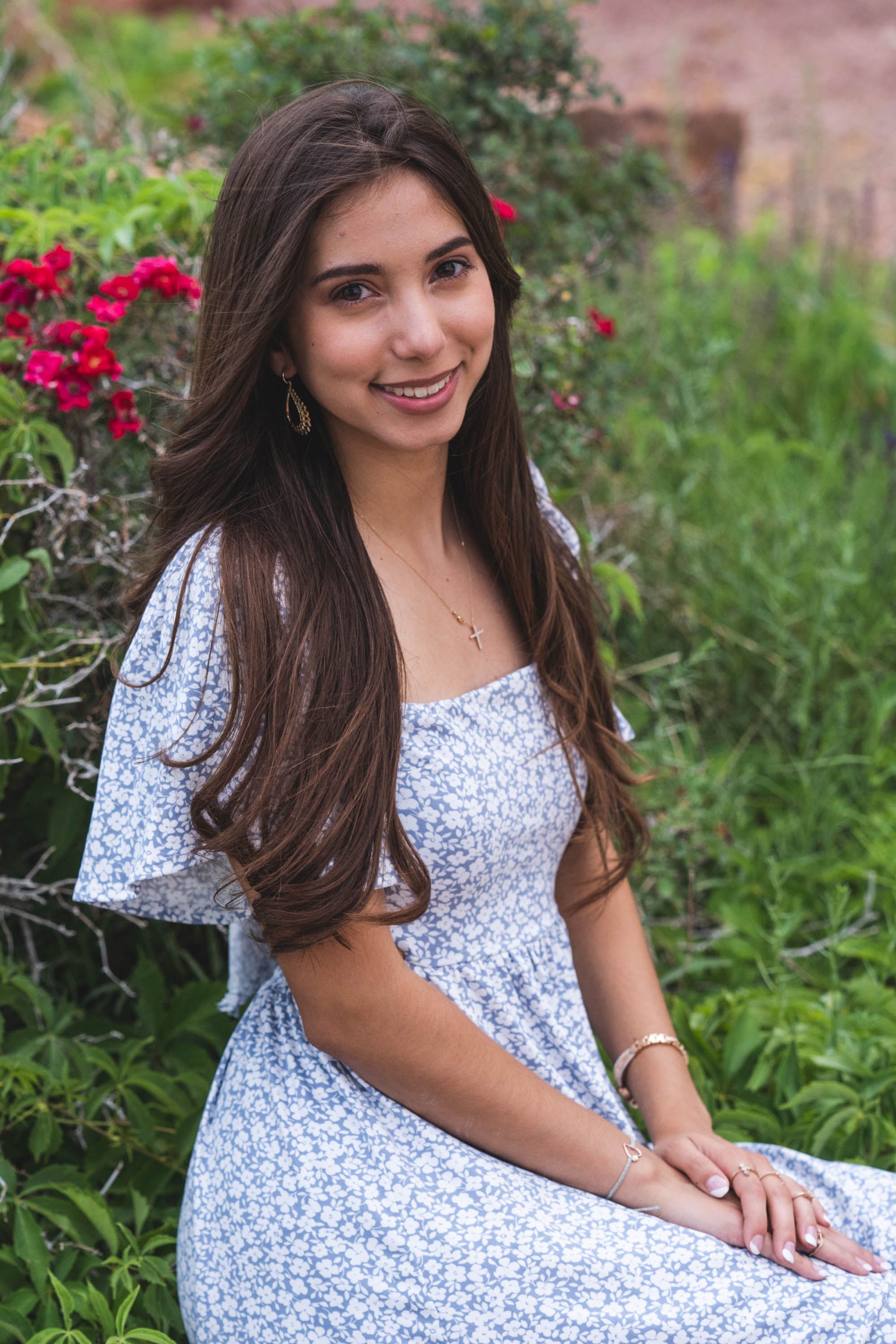 Young woman in floral dress posing by red flowers in garden setting.