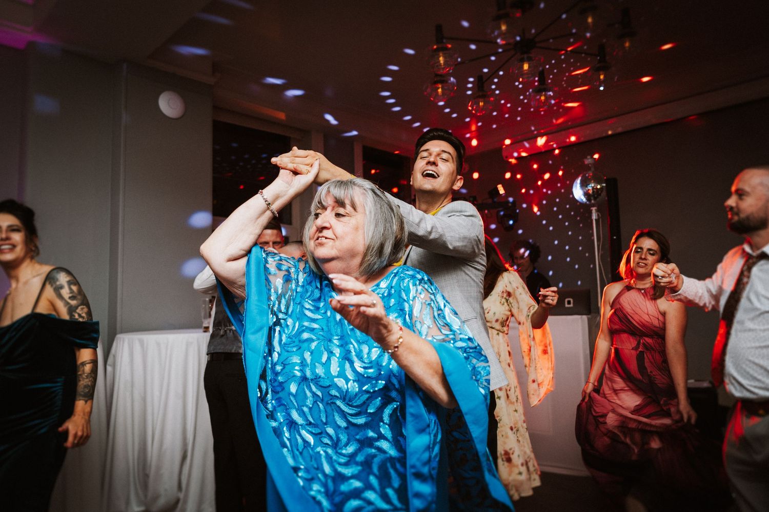 People dancing and celebrating at a lively party with red lighting and disco ball reflections on the dance floor.