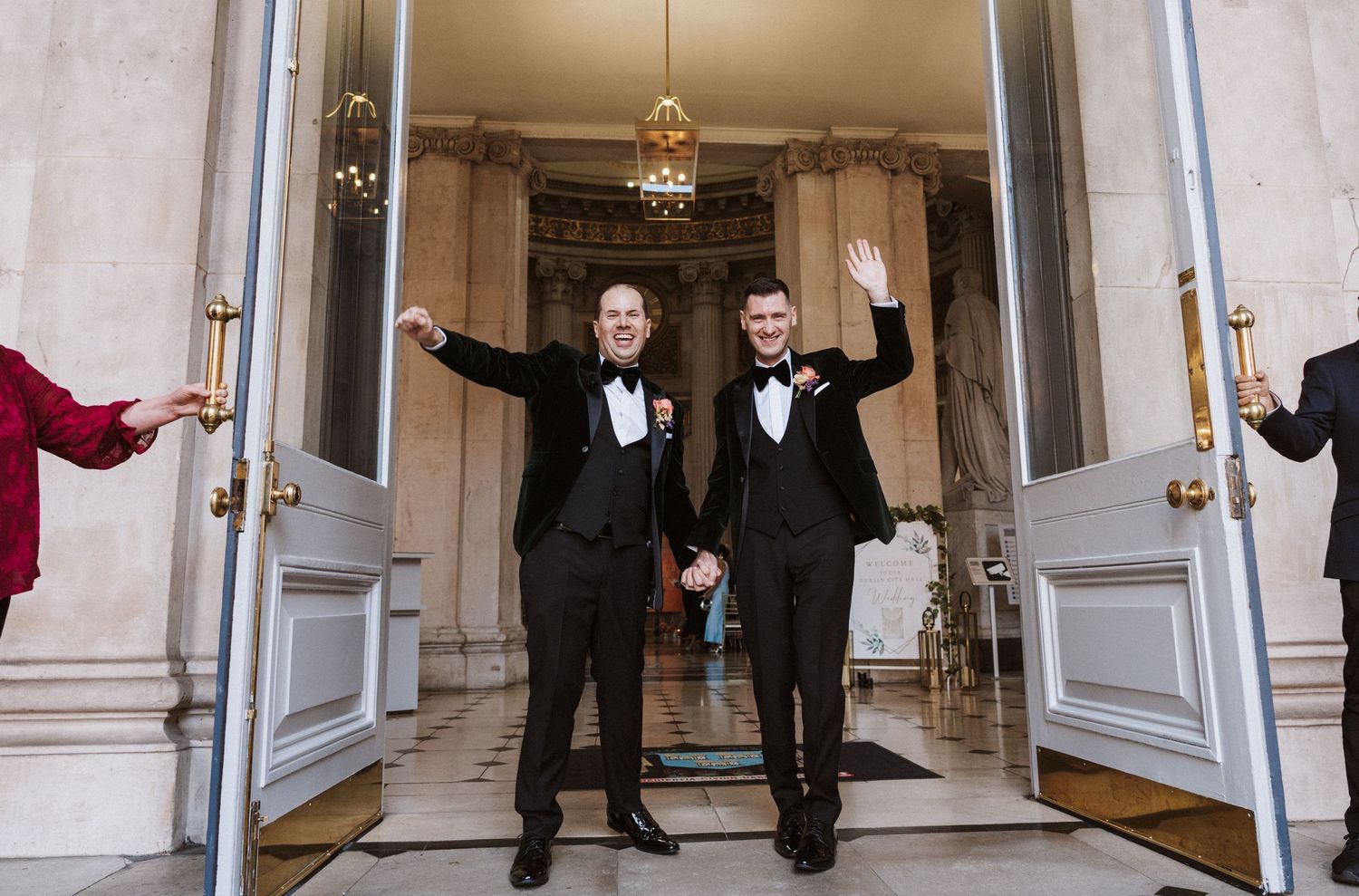 Two people in tuxedos leap joyfully down an elegant hallway with white walls and ornate decor.