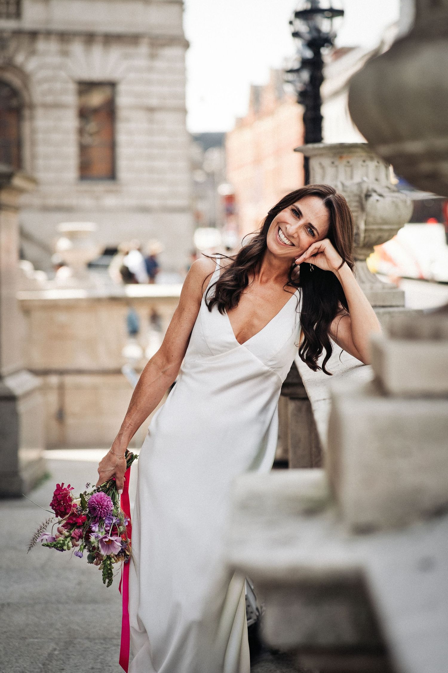 Smiling bride in white satin gown holding colorful bouquet with pink ribbon, posing outdoors near stone architecture.
