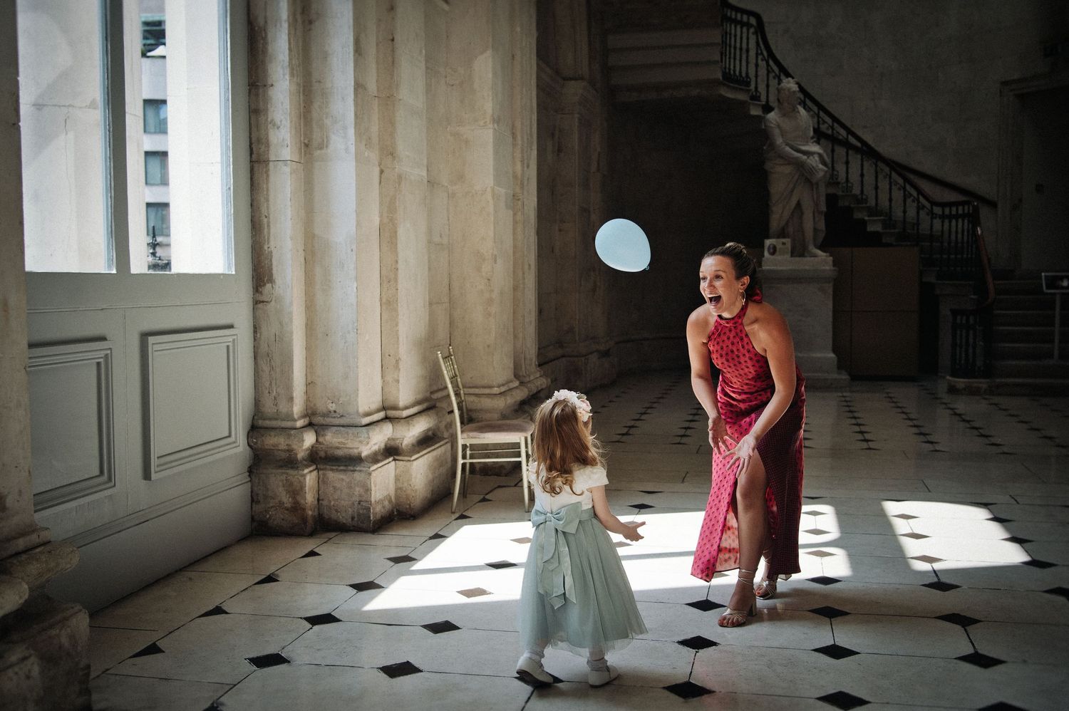 Woman in red dress and little girl in blue tutu playing with a balloon in an elegant marble hallway.