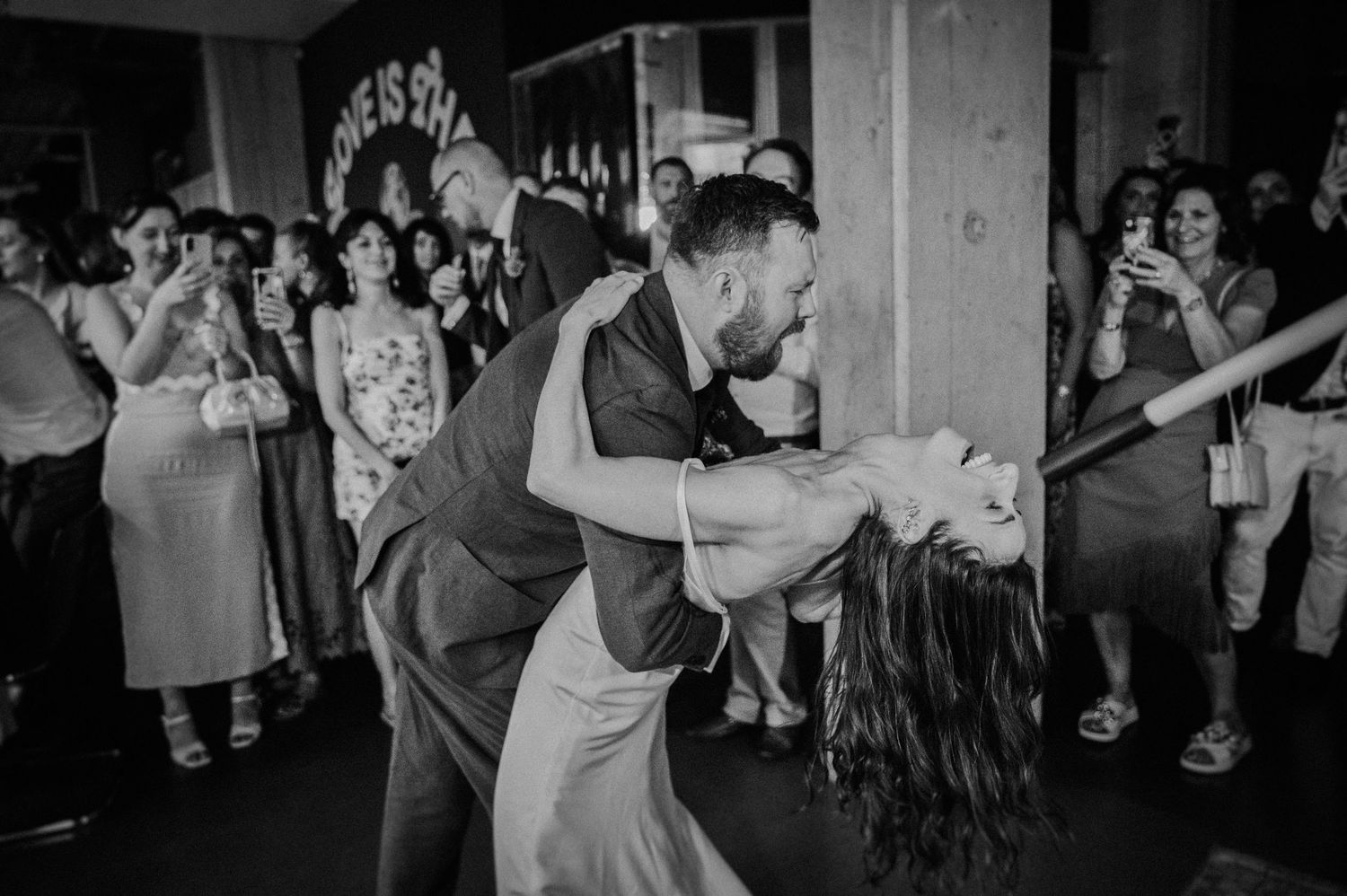 Groom dips bride dramatically on dance floor surrounded by cheering wedding guests in black and white photo.