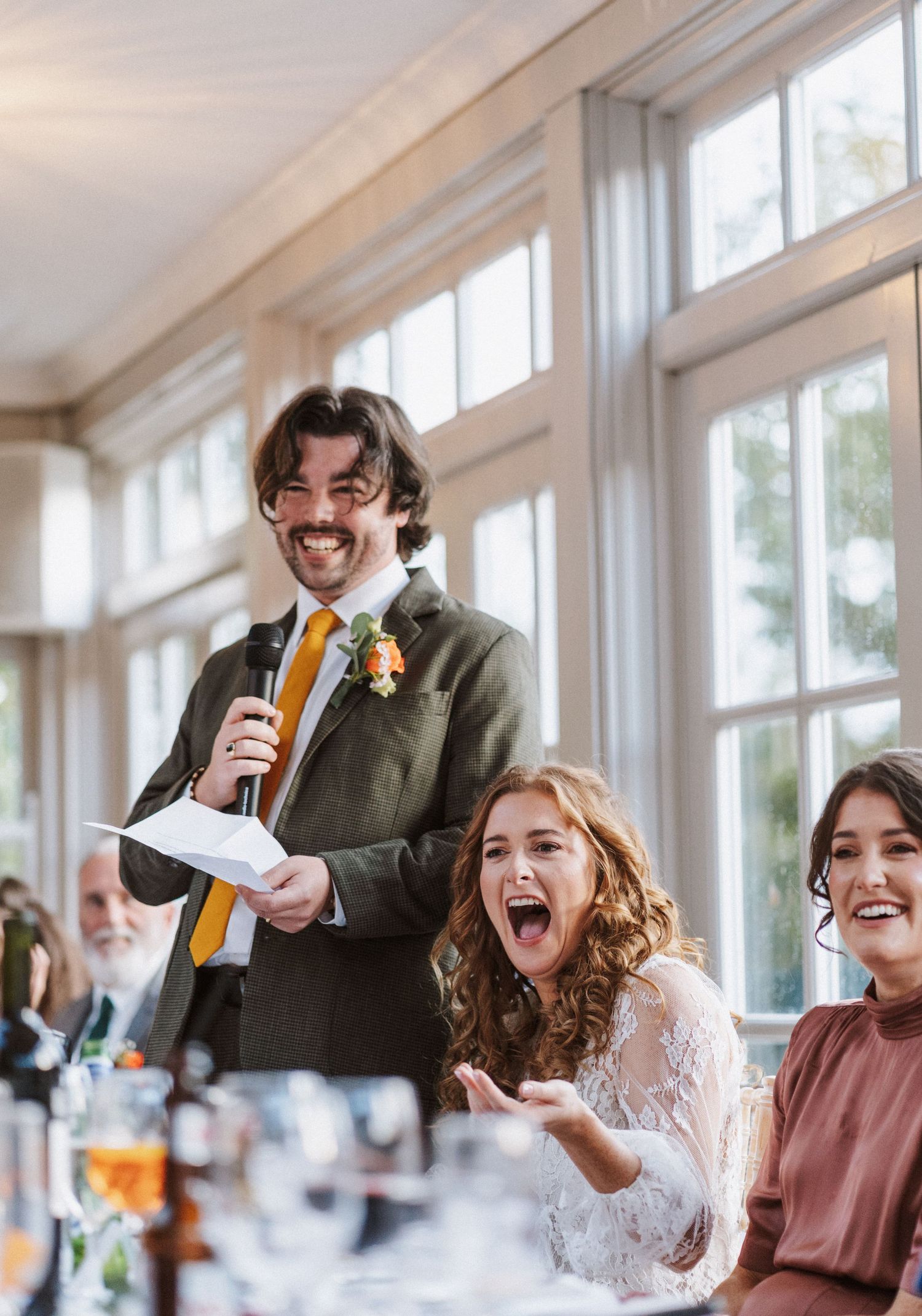 A joyful wedding reception scene with laughter and celebration at a bright venue with large windows.