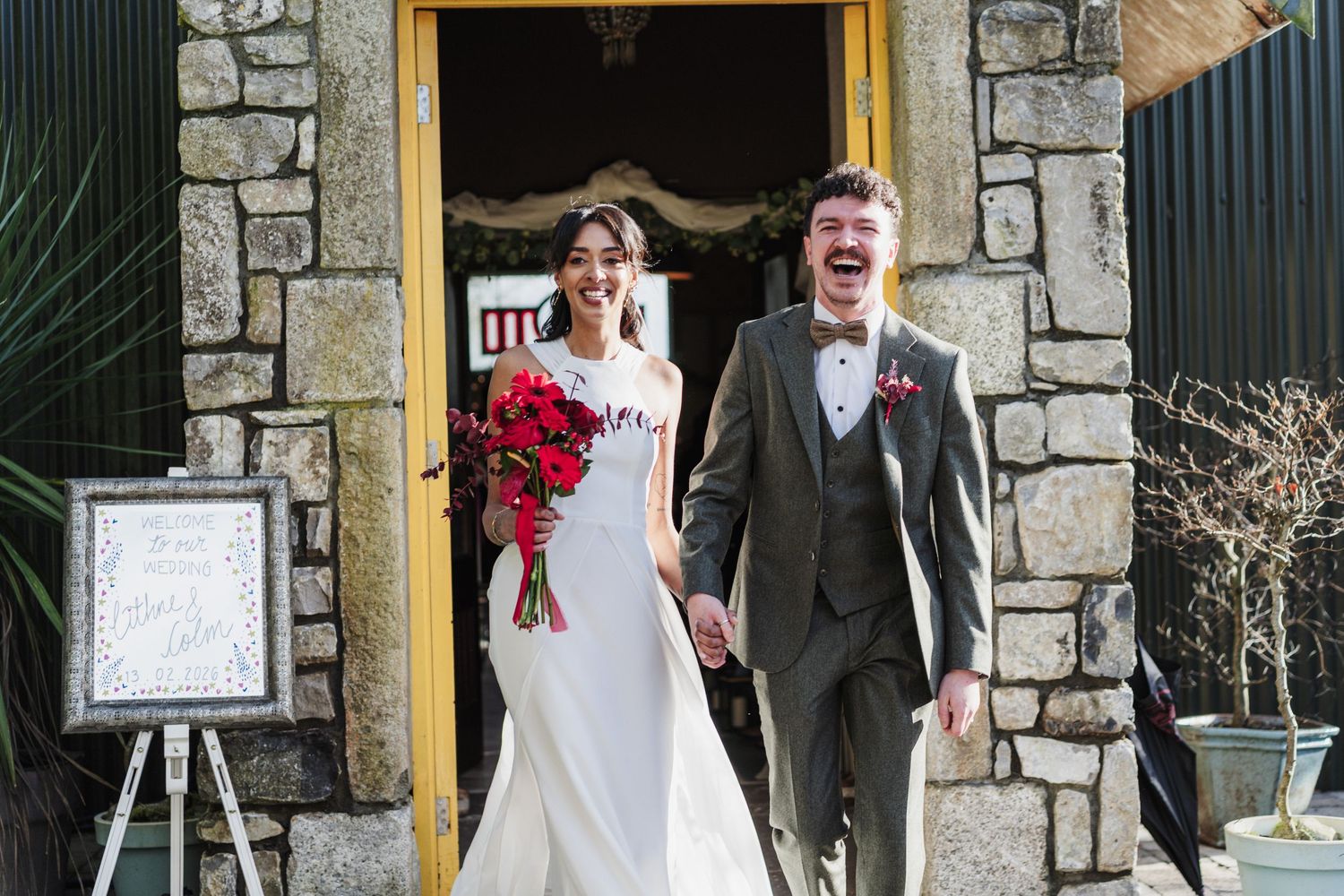 A joyful couple exits a stone building on their wedding day, with the bride holding a vibrant red bouquet.