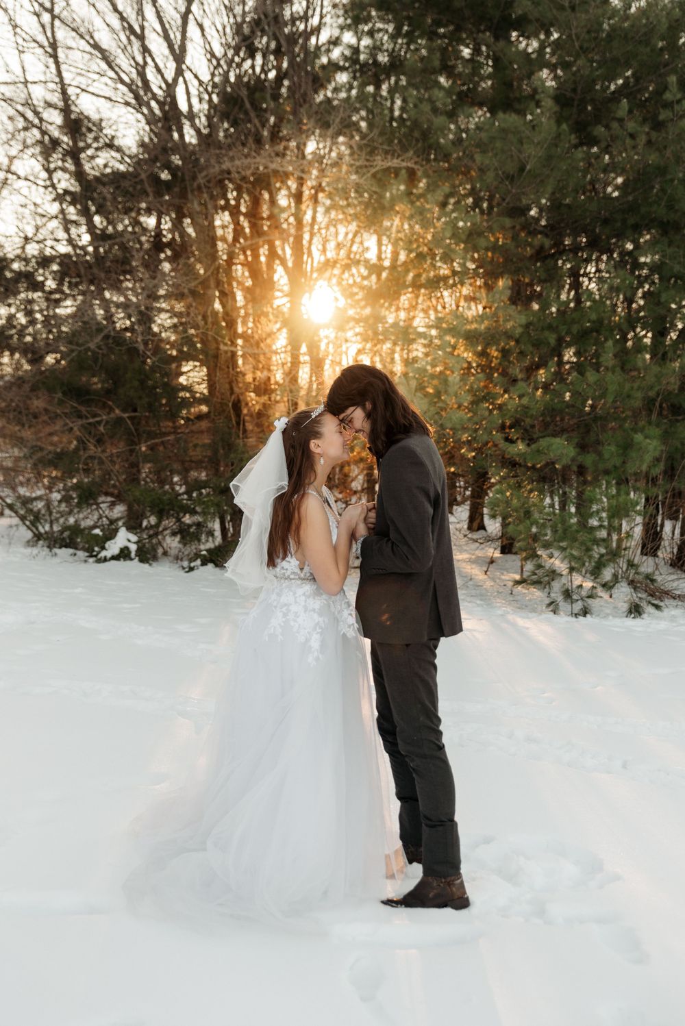 Couple takes romantic winter wedding photos in snowy forest setting at sunset.