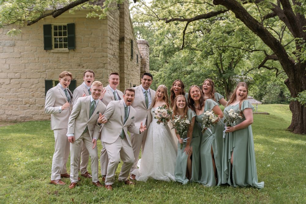 Wedding party poses outdoors in sage green bridesmaid dresses and tan suits near a stone building.