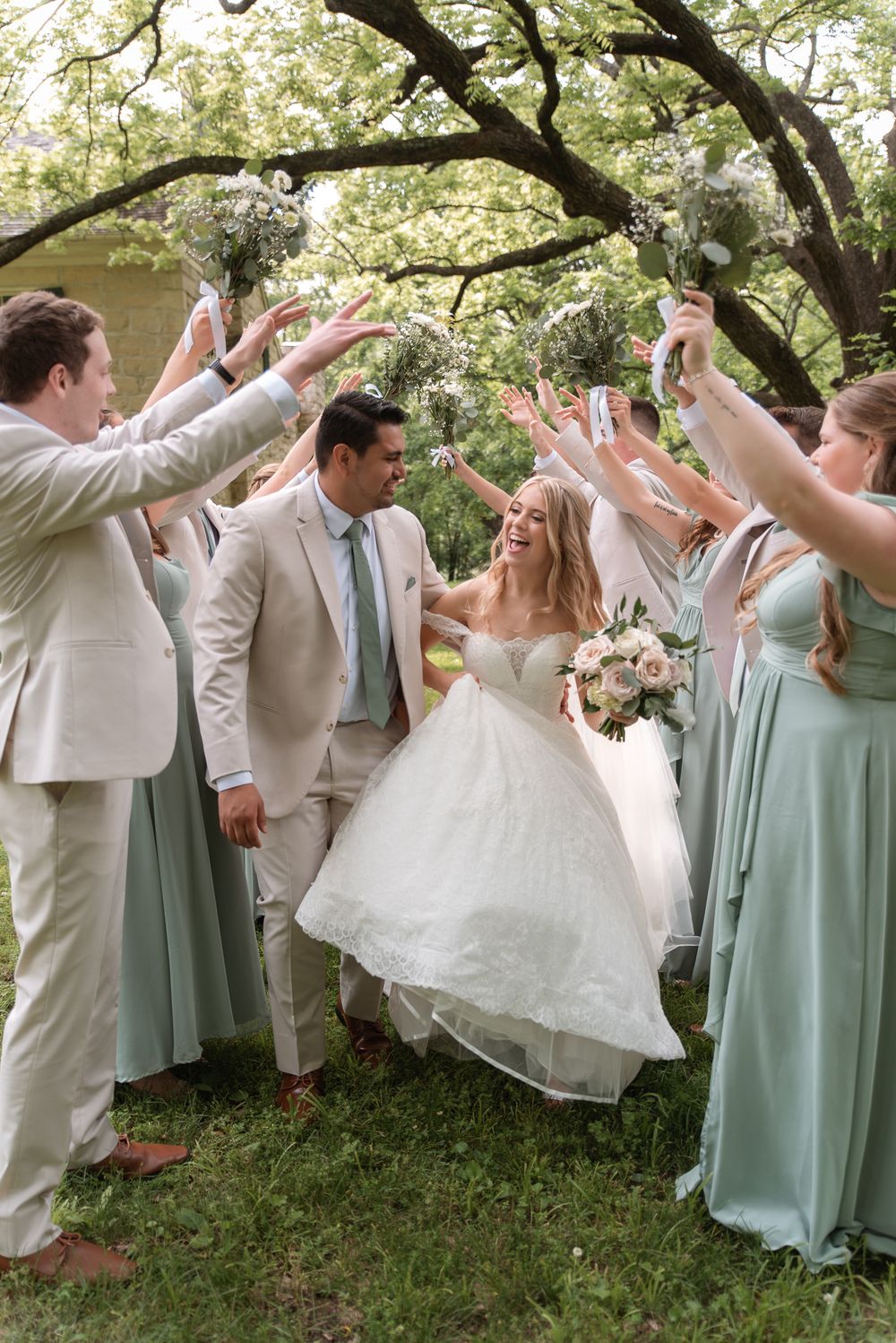 Bride and groom walk through arch of raised hands with wedding party in sage green dresses and tan suits outdoors.