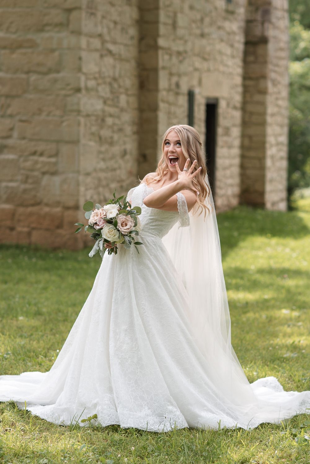 Excited bride in white off-shoulder gown with cape train holds bouquet outdoors near stone building.