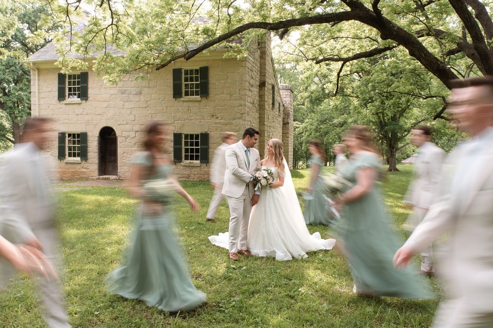 Bride and groom share a romantic moment outdoors as blurred bridesmaids in sage green gowns move around them.