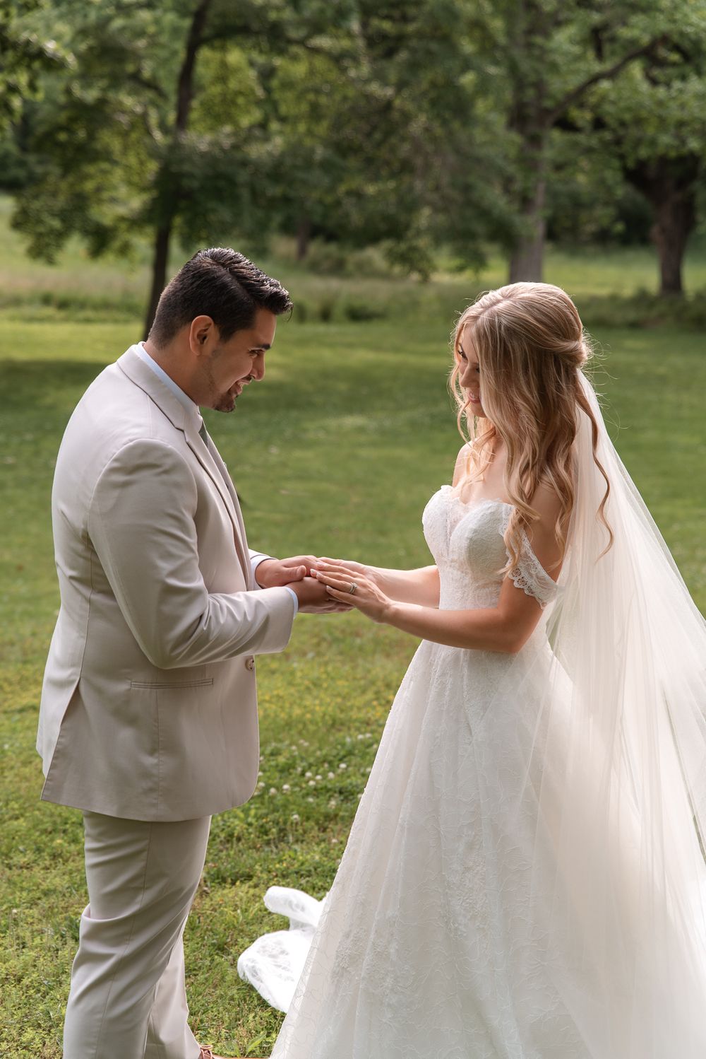 Bride and groom exchange rings outdoors in lush green garden, bride in lace gown with veil, groom in beige suit.