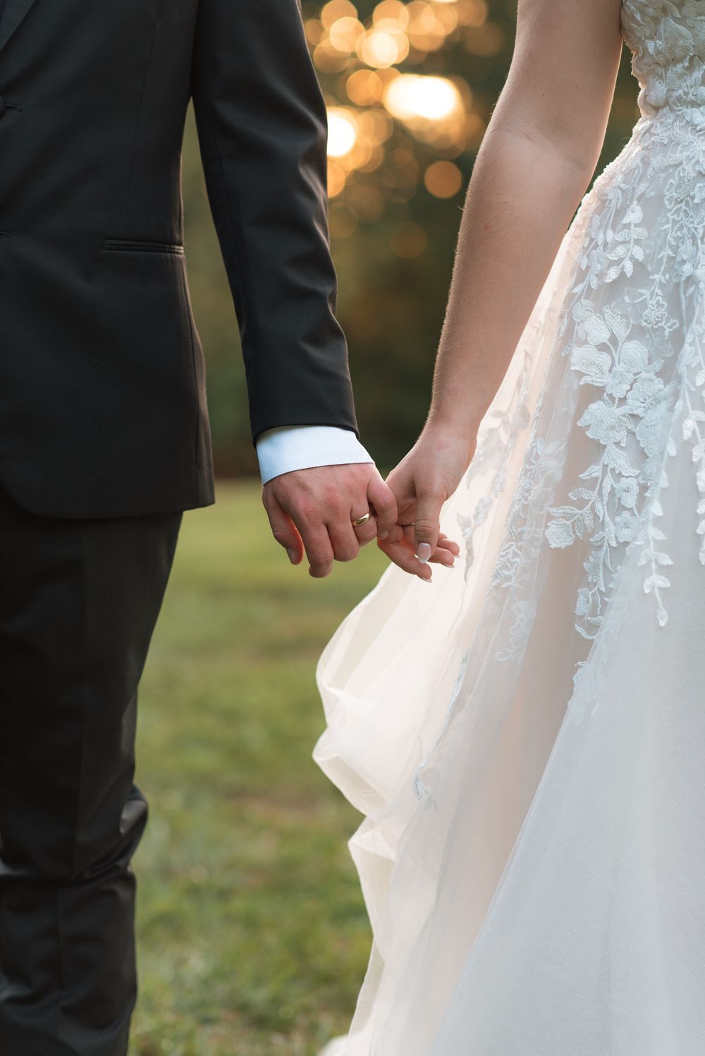 Bride and groom holding hands outdoors, wearing a white lace wedding gown and dark suit, with bokeh lights in background.