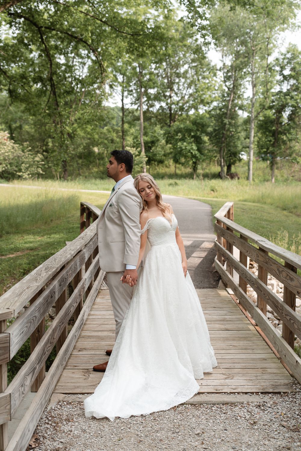 Bride in white gown and groom in gray suit hold hands on a wooden bridge surrounded by lush green trees.
