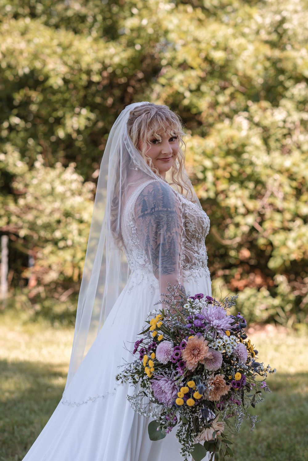 Bride in white gown and veil holds colorful wildflower bouquet outdoors, showcasing tattoo sleeves and blonde hair.