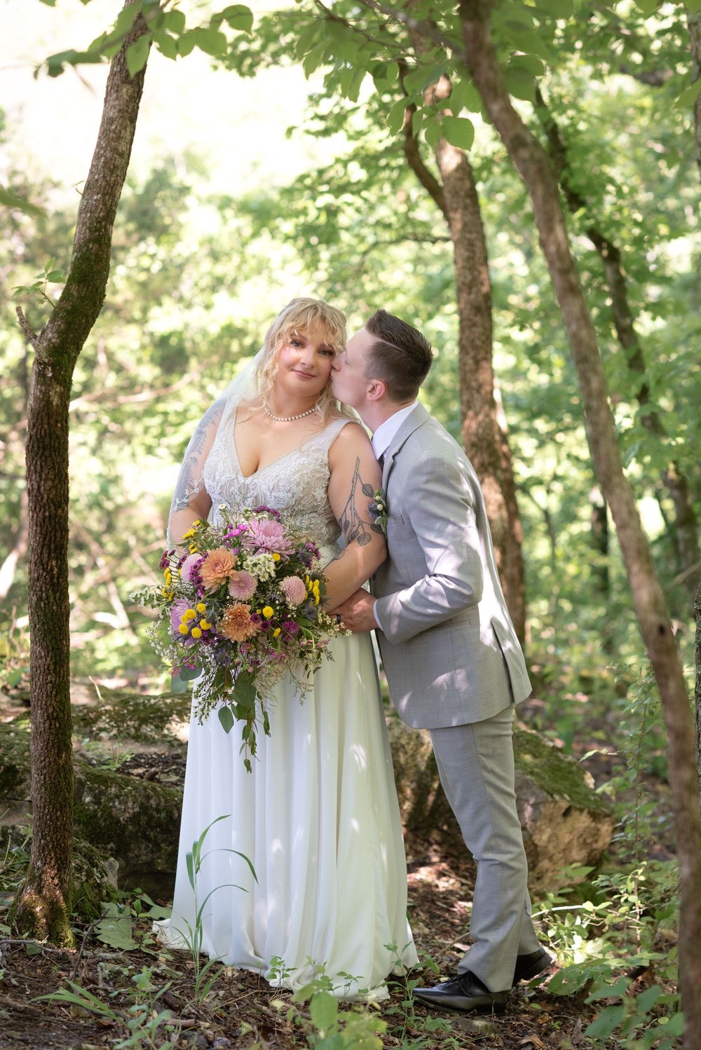 Bride holding colorful bouquet receives kiss from groom in gray suit amid lush green forest wedding setting.