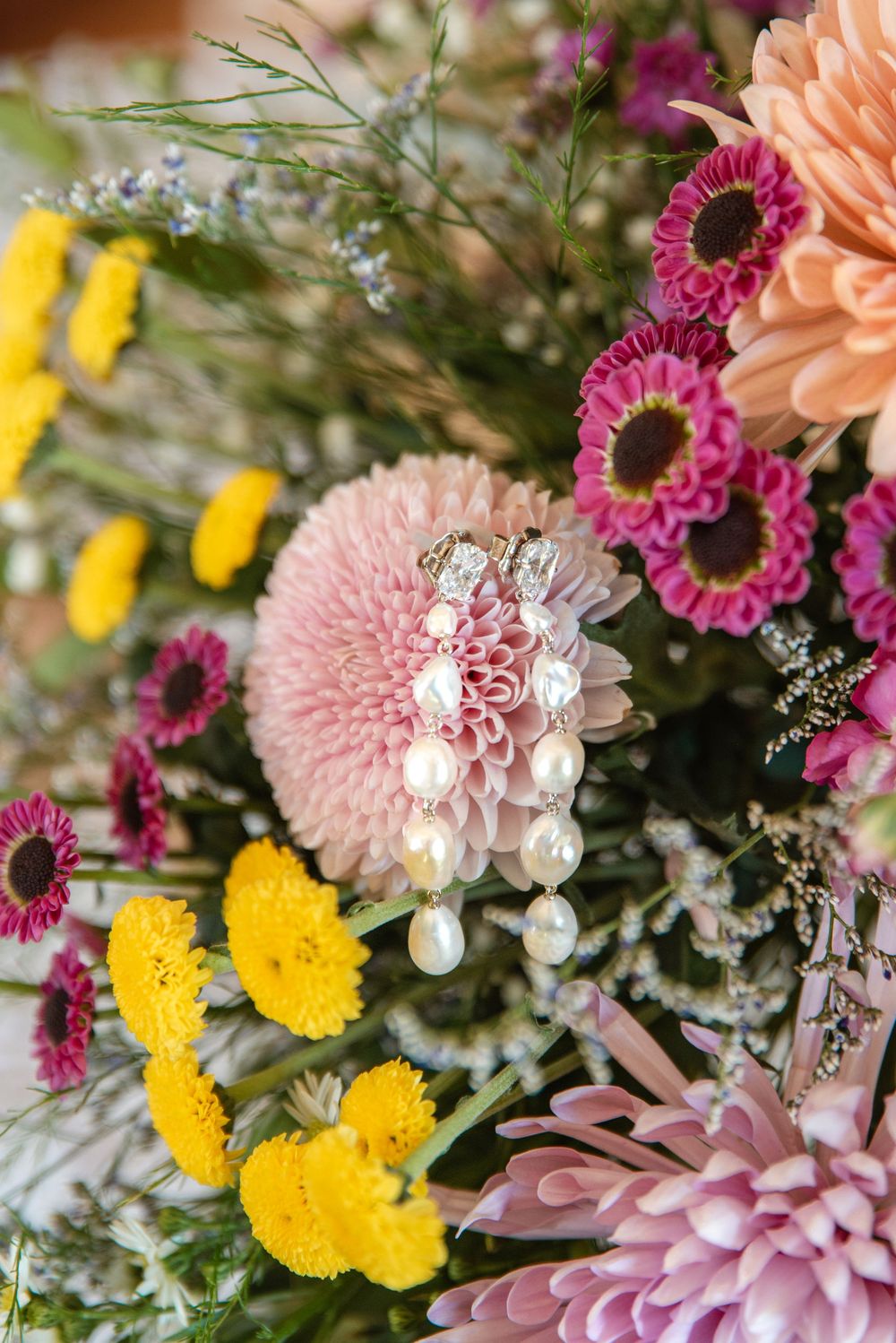 Pearl drop earrings resting on a pink dahlia surrounded by colorful yellow, magenta, and peach flowers in a bridal bouquet.
